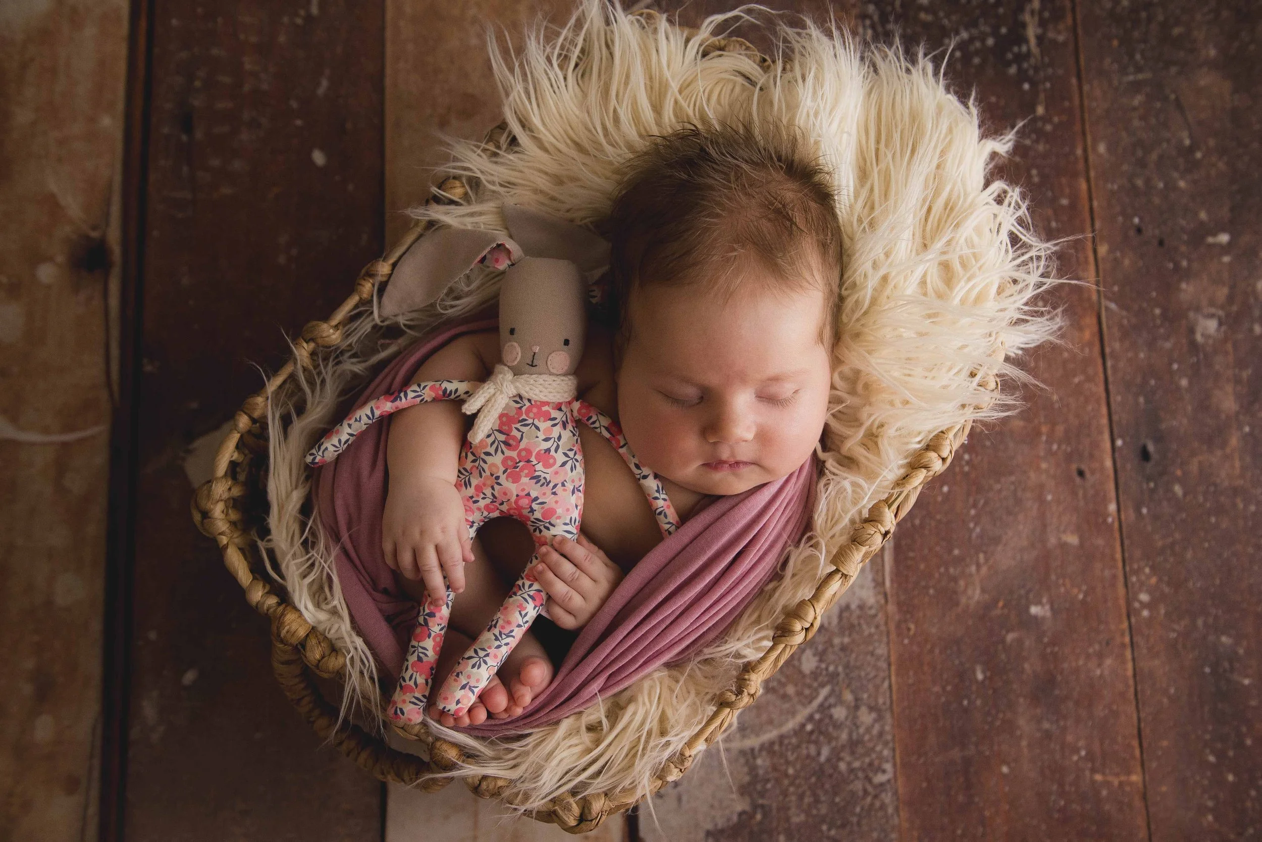A sleeping baby girl with brown hair and closed eyes, wrapped in pink cloth, lying in a wicker basket on a wooden floor, with a small plush bunny toy resting on her chest.