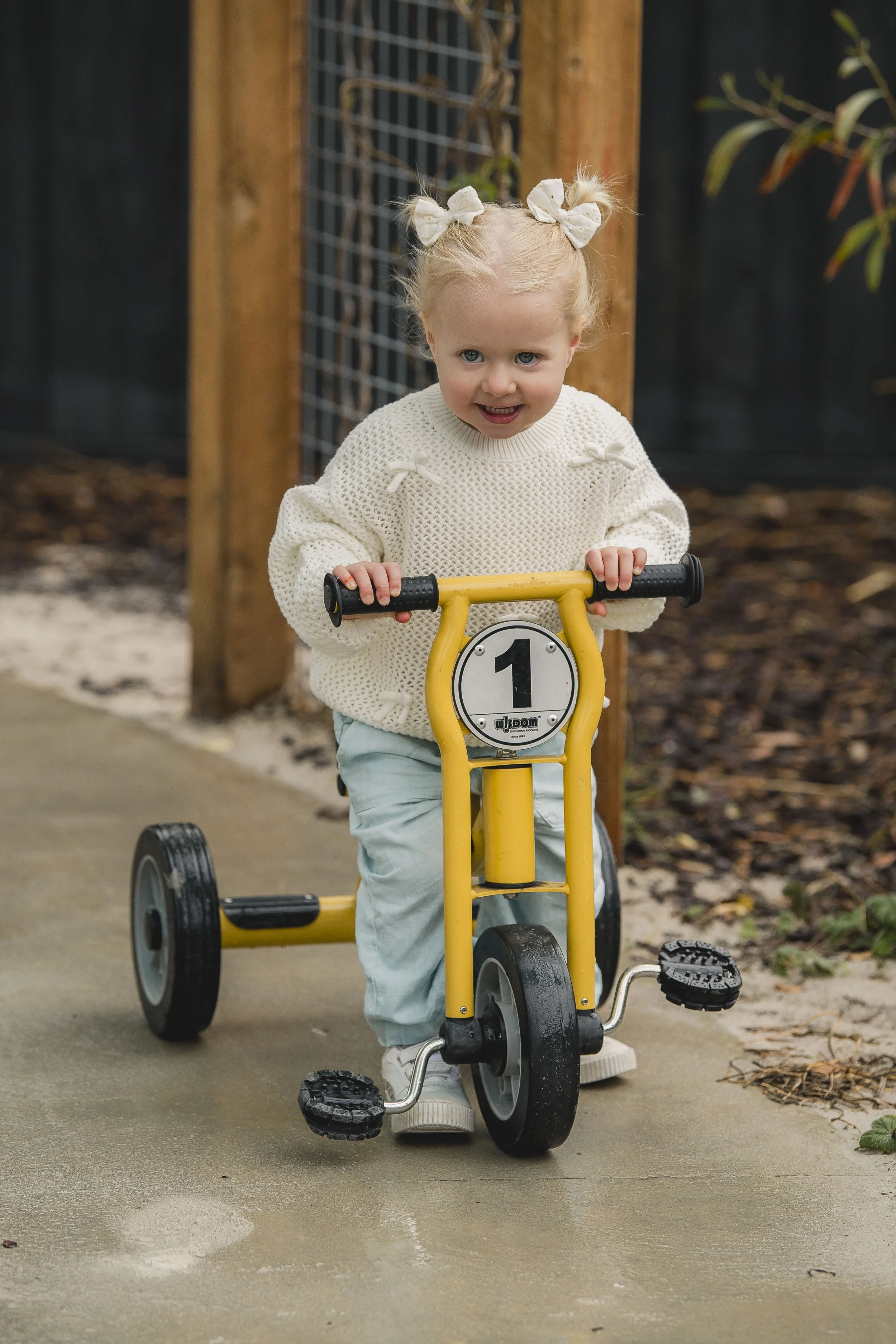 A young girl with blonde hair tied in two pigtails with bows, riding a yellow tricycle on a sidewalk in a backyard. She is wearing a white knitted sweater and light-colored pants. The background includes a wooden fence and some foliage.