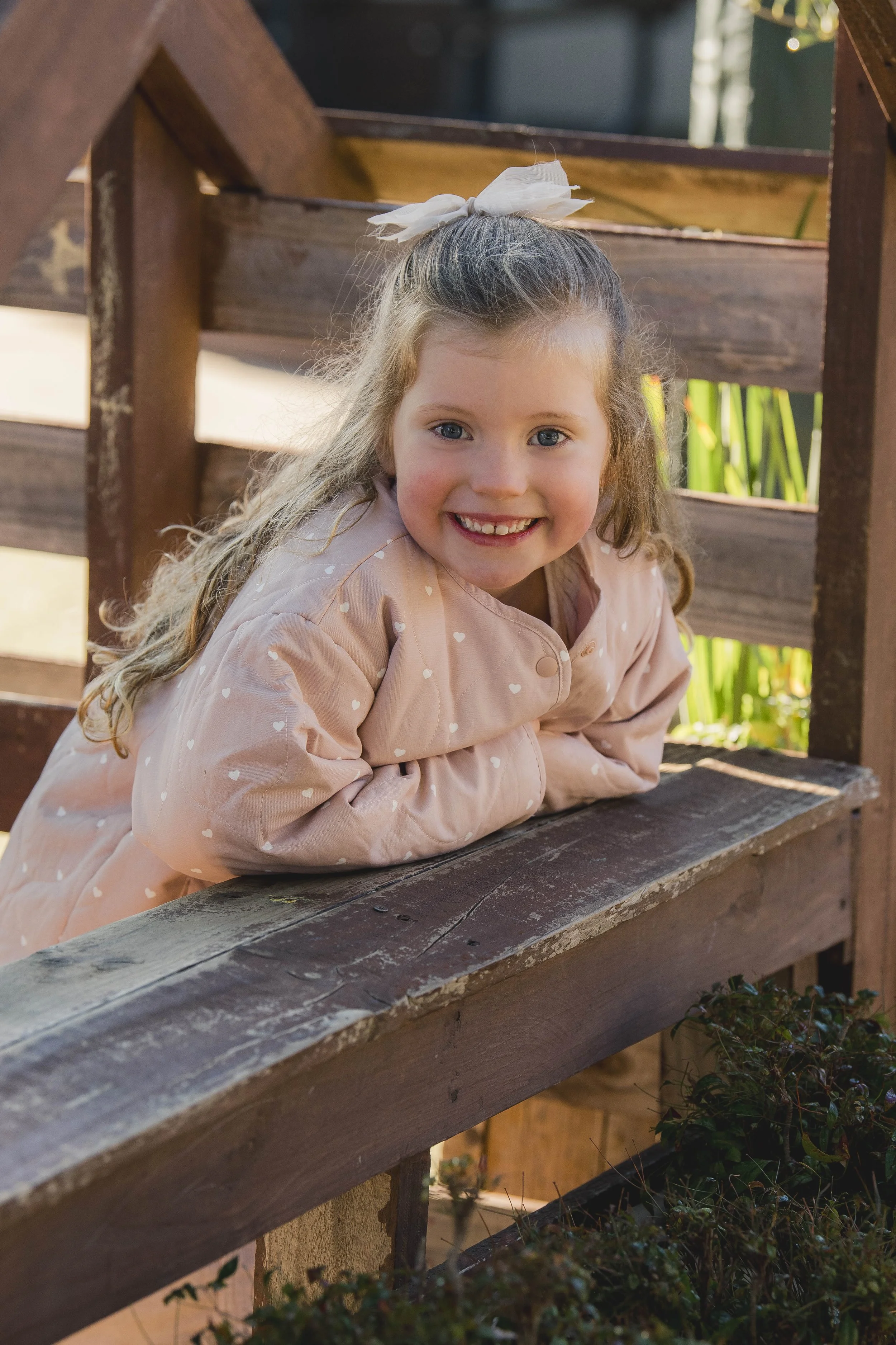 A young girl with long curly hair, smiling and leaning on a wooden railing outdoors on a sunny day, wearing a light pink jacket with white hearts and a white bow in her hair.
