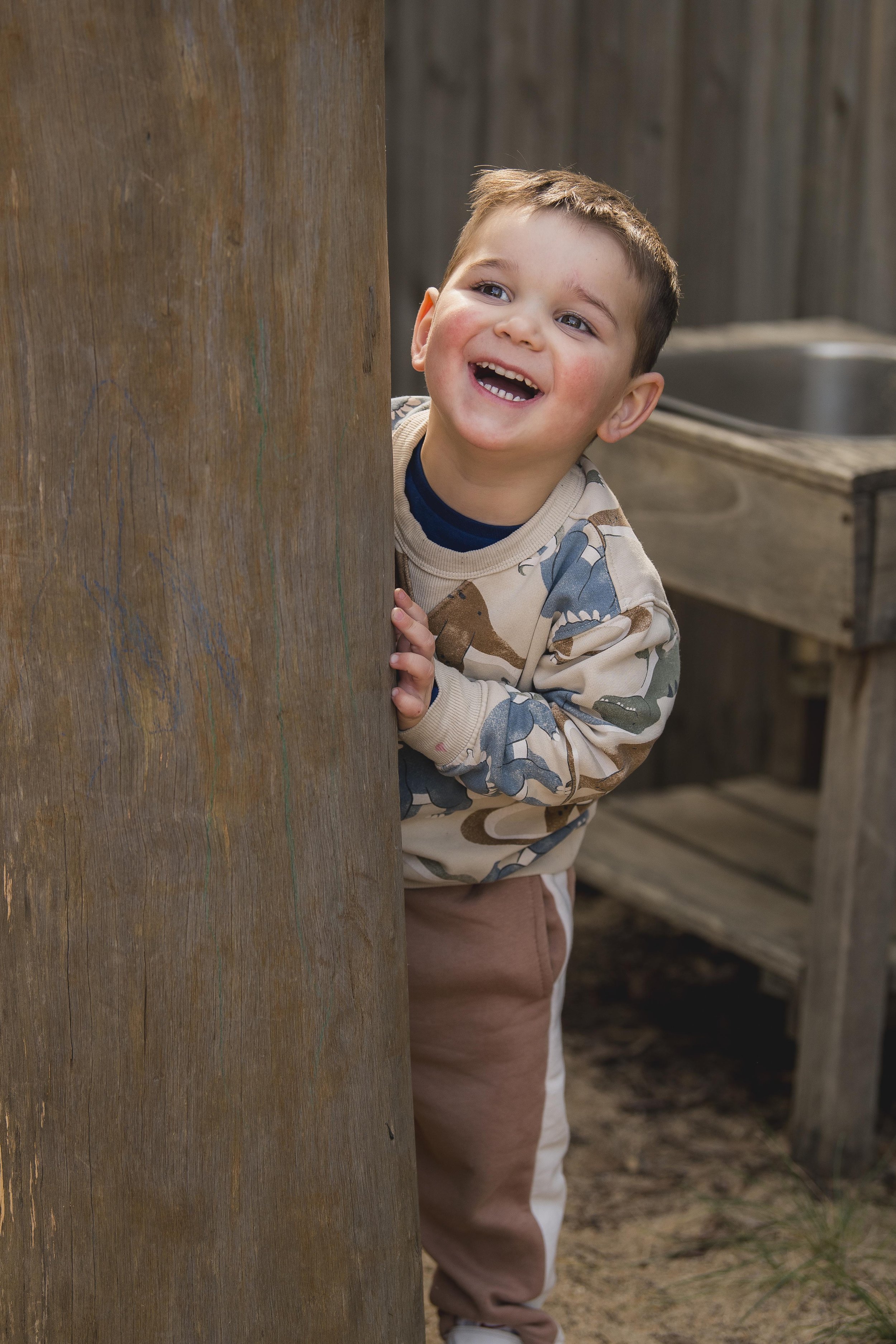 A young boy with short brown hair and blue eyes peeking from behind a wooden post, smiling with his mouth open, wearing a camouflage-patterned long-sleeve shirt and beige pants, in a rustic outdoor setting with a wooden sink and dirt ground.
