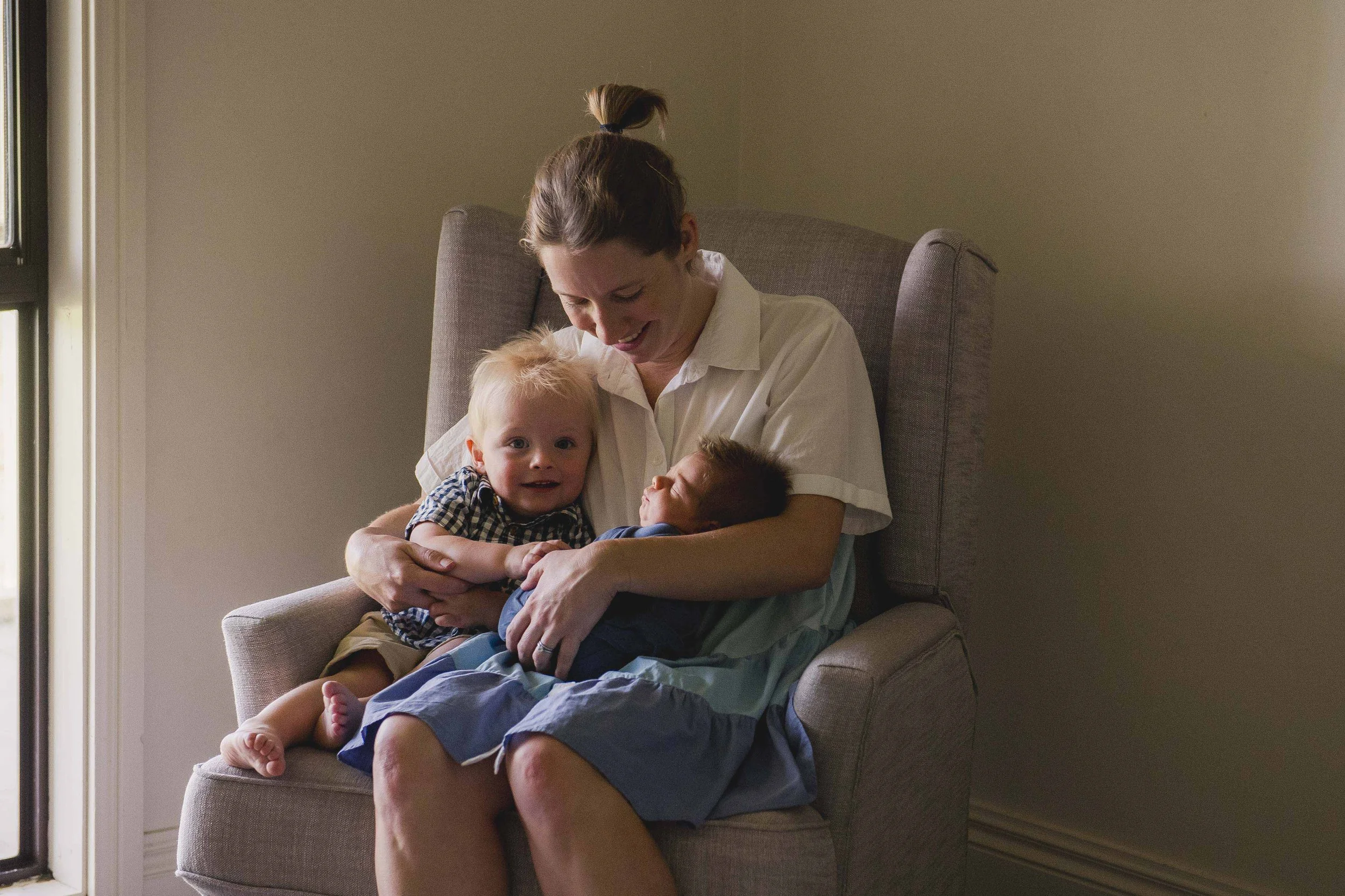 A woman sits on a gray armchair, holding and cuddling two young children, one with blond hair and one with brown hair. They are indoors next to a window, with beige walls.