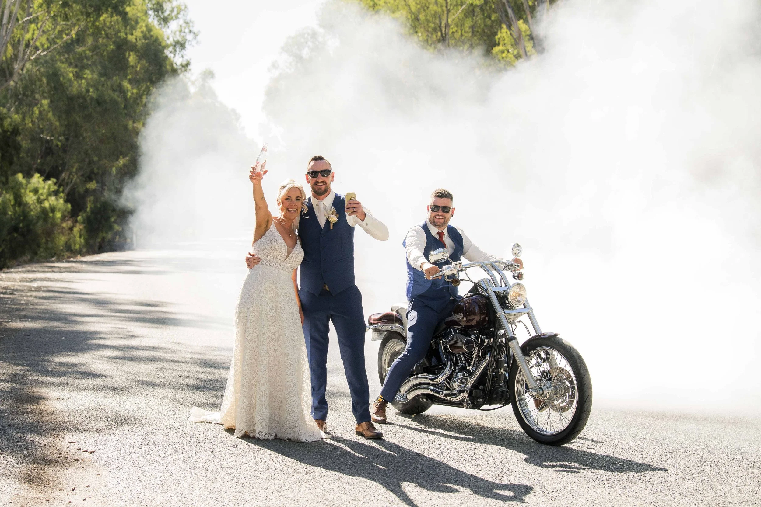 A bride and groom standing next to a man on a motorcycle on a dusty road surrounded by trees, with the bride holding a drink and the groom raising a glass, all smiling.