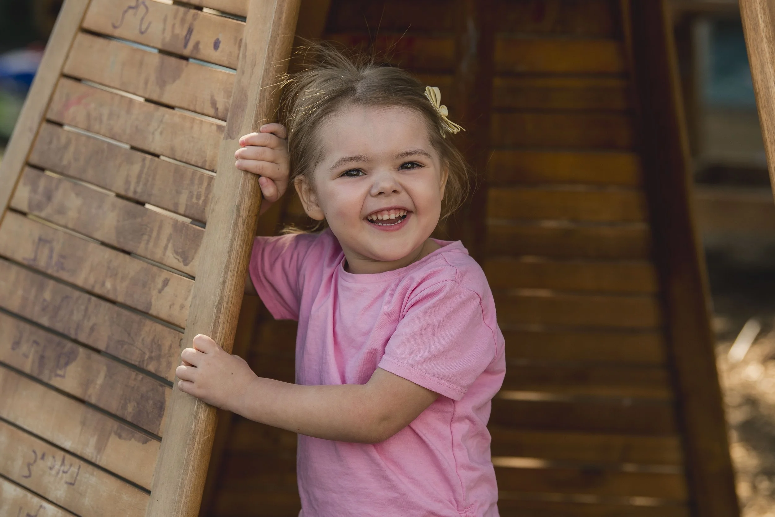 A young girl with brown hair, a yellow bow, and a pink shirt smiling and playing on a wooden playground structure.