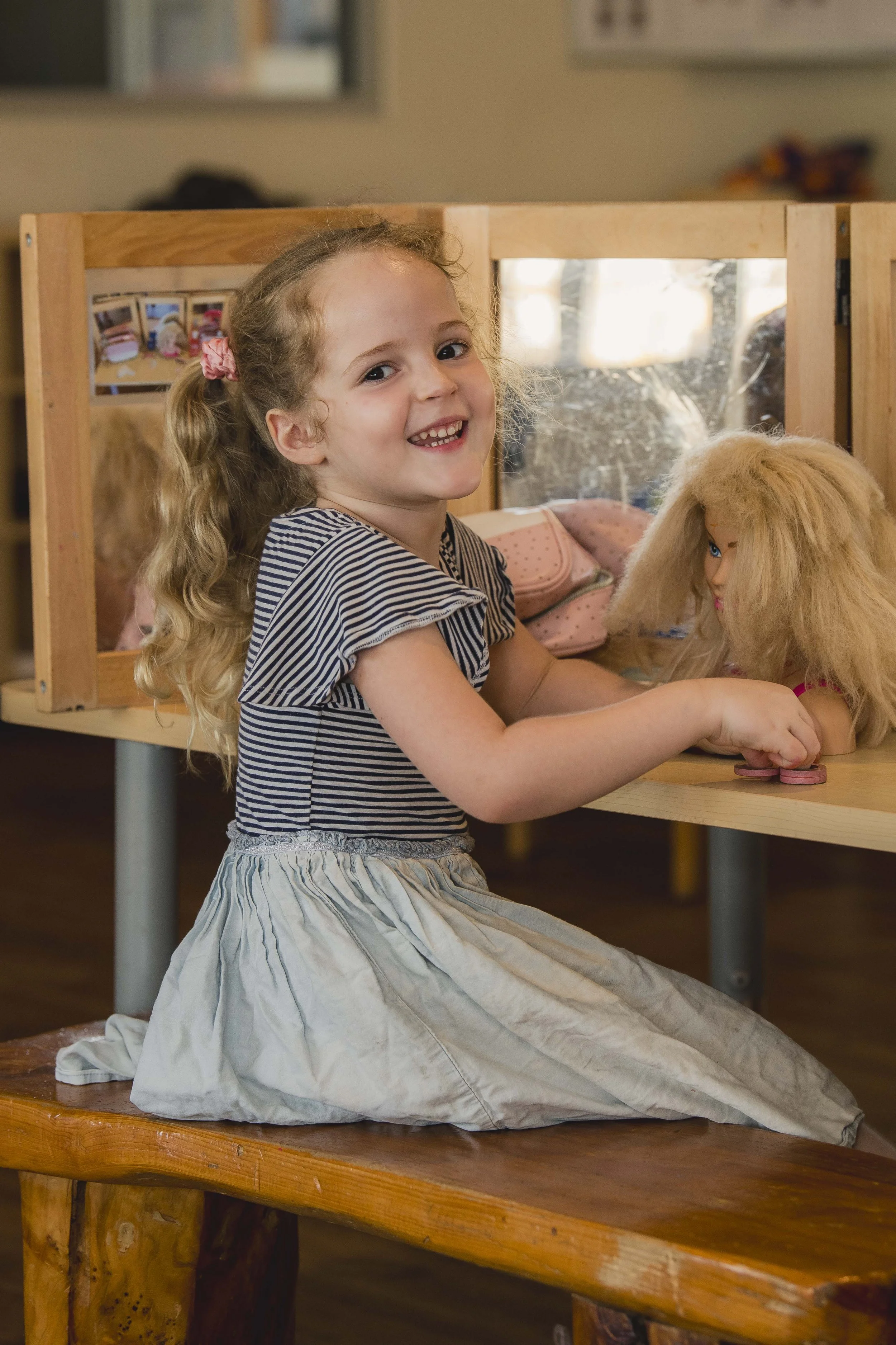 A young girl with curly blonde hair and a pink hair tie, smiling, sitting on the floor in front of a puppet theater with a doll. She is wearing a striped top and a light-colored skirt, with a wooden table in front of her.