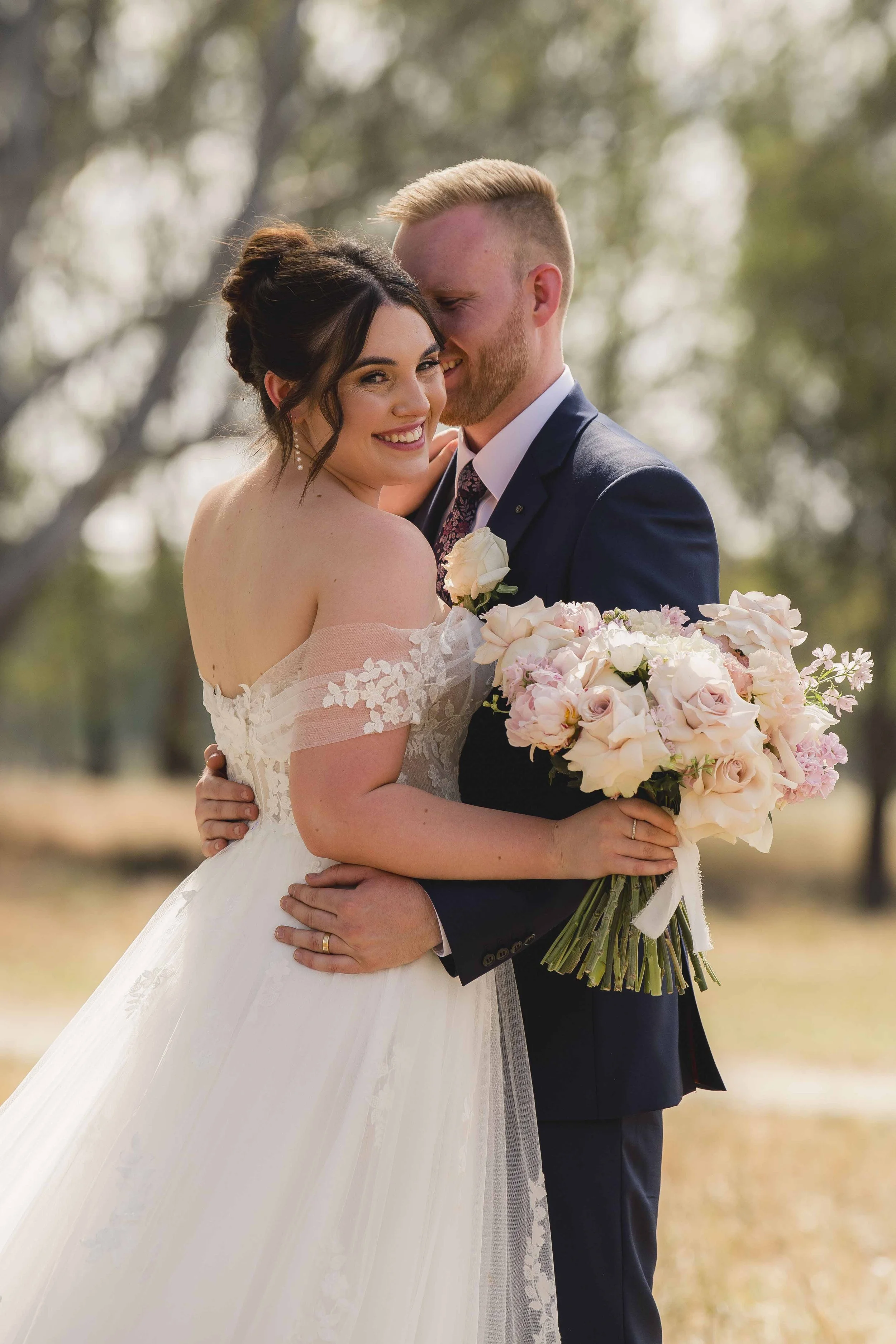 A bride and groom embrace outdoors during daytime, the bride holding a bouquet of pink and white flowers, both smiling and close together.