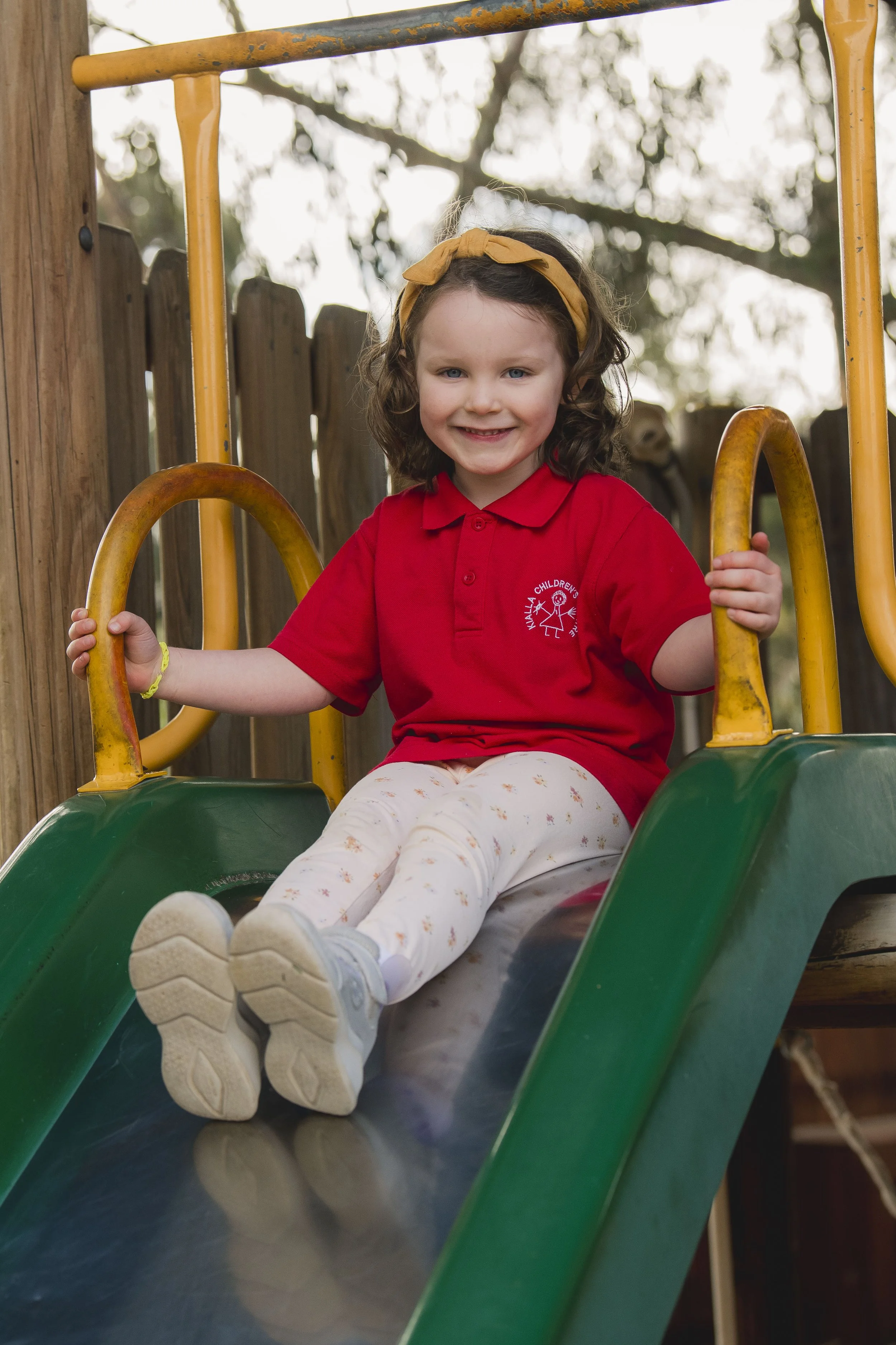 Young girl with curly hair, wearing a yellow headband and red shirt, sitting on a slide in a playground, smiling at the camera.