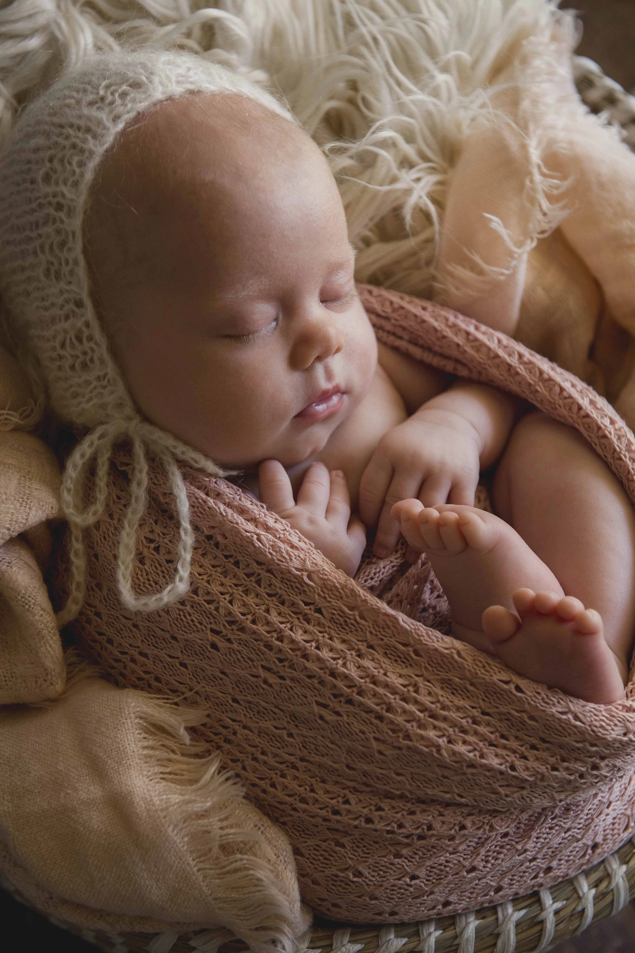 A sleeping baby with a headband, wrapped in a pink knitted blanket, lying on a woven basket.