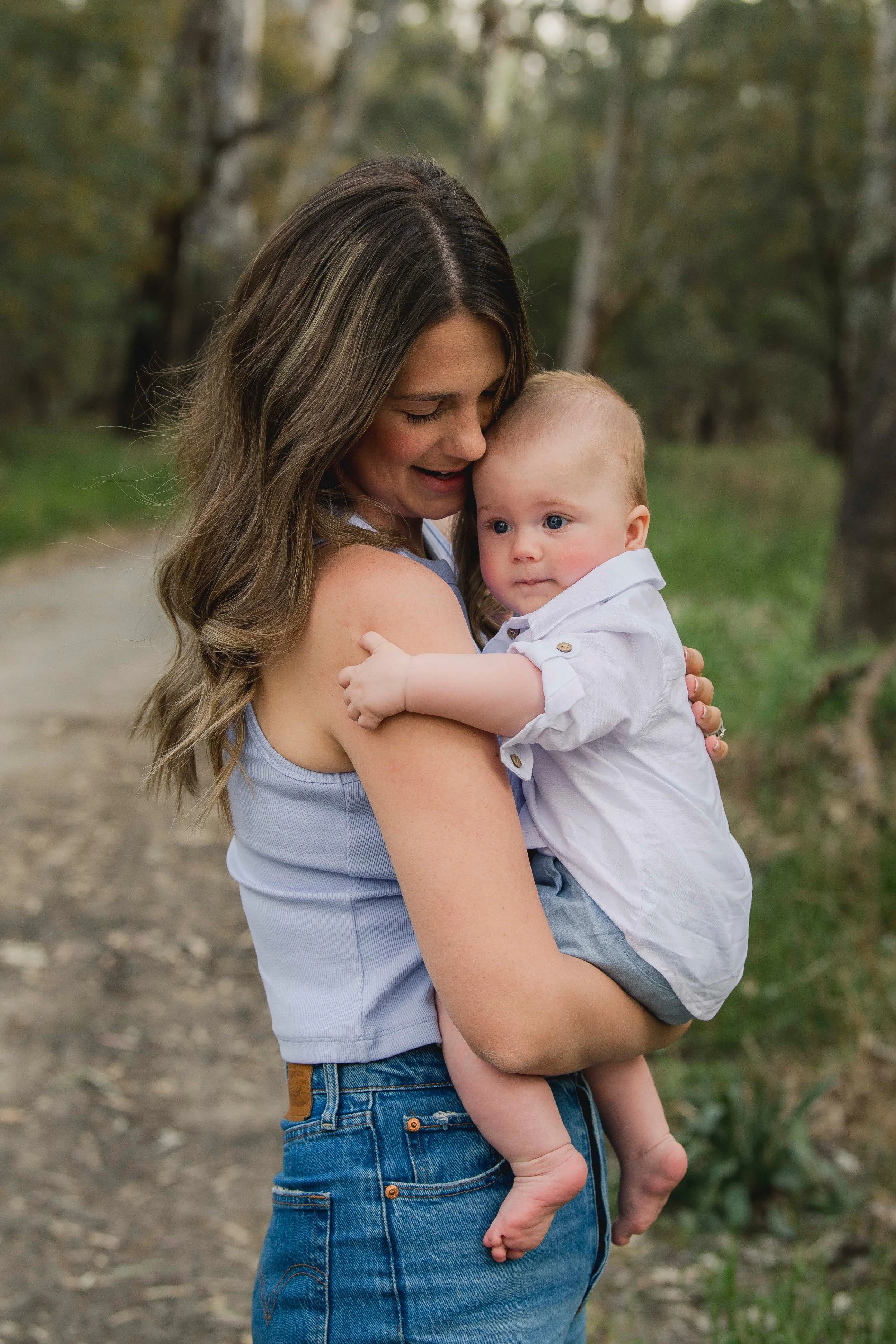 A woman holding a young child outdoors on a dirt path with trees in the background.