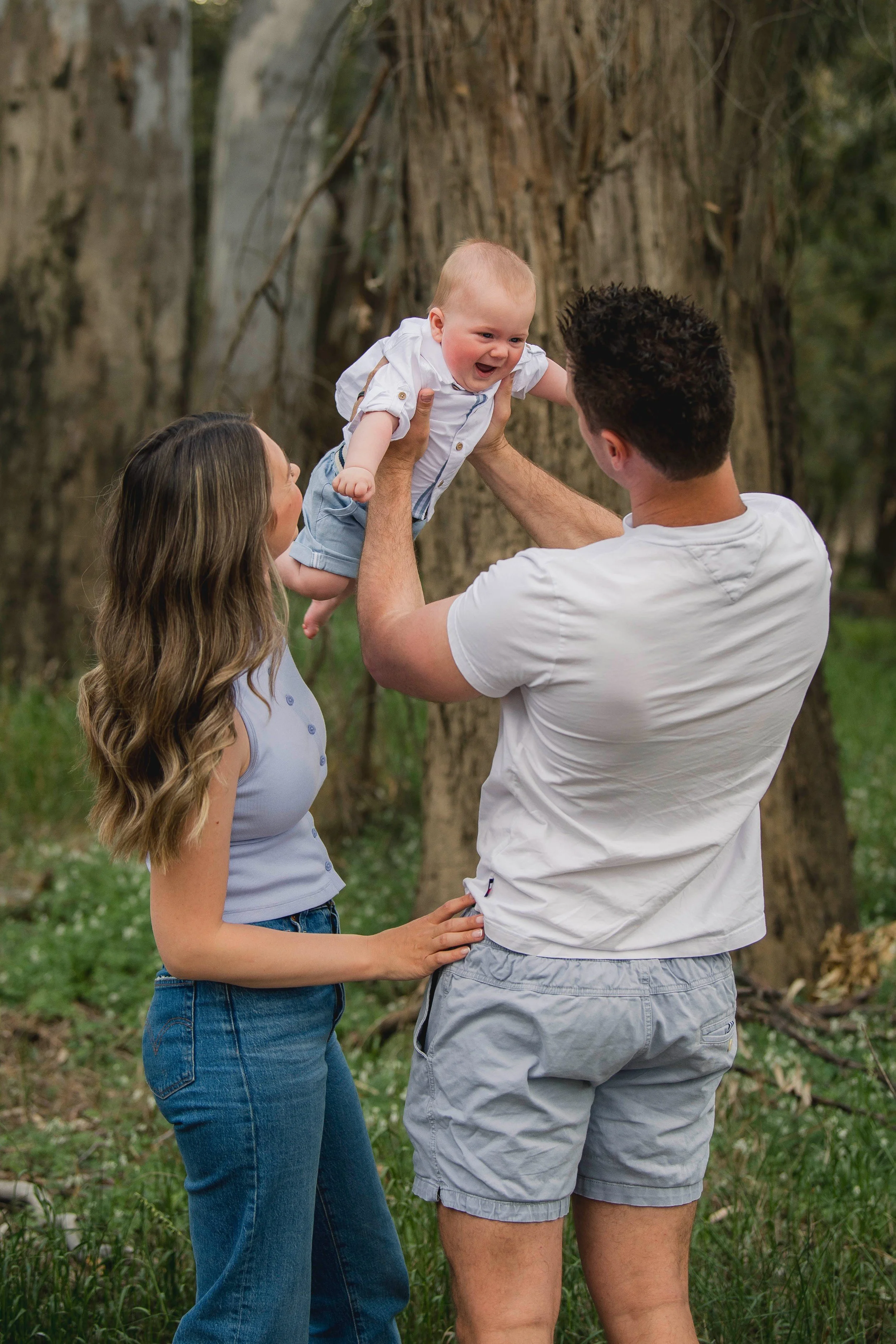 A family of three outdoors near a large tree; a man and woman are holding a smiling baby in the air between them. The woman has long wavy hair and is wearing a sleeveless top, the man has short dark hair and is wearing a white t-shirt, and the baby i