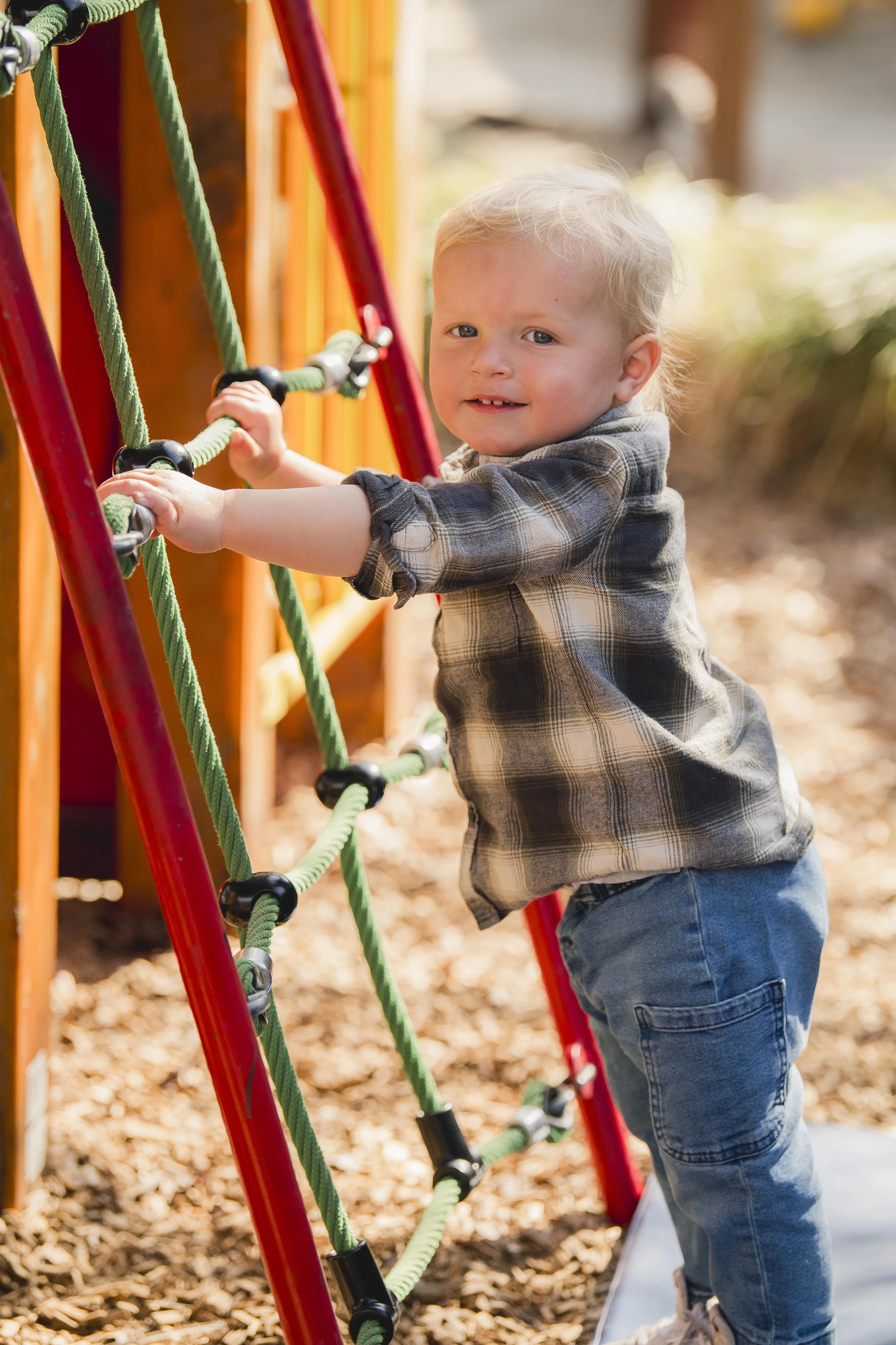 A young boy with blond hair, wearing a gray plaid shirt and jeans, playing on a jungle gym with green ropes outdoors on a sunny day.