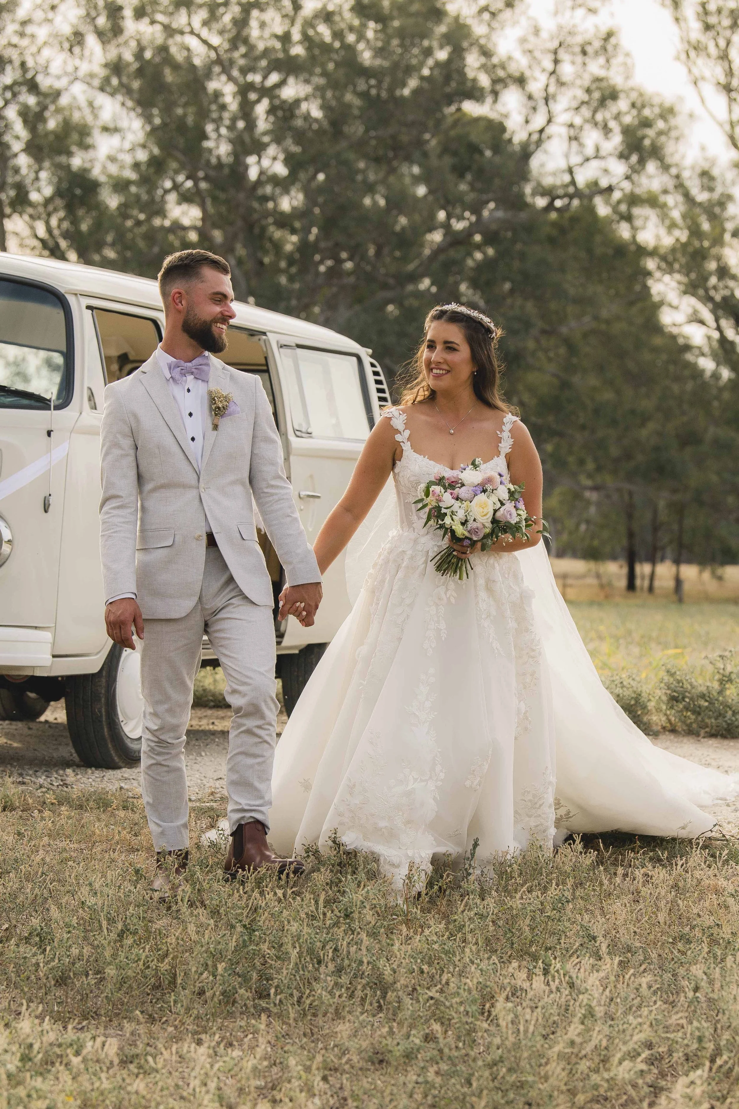 A bride and groom walking hand in hand outdoors near a vintage white van, surrounded by trees and grass, during sunset.