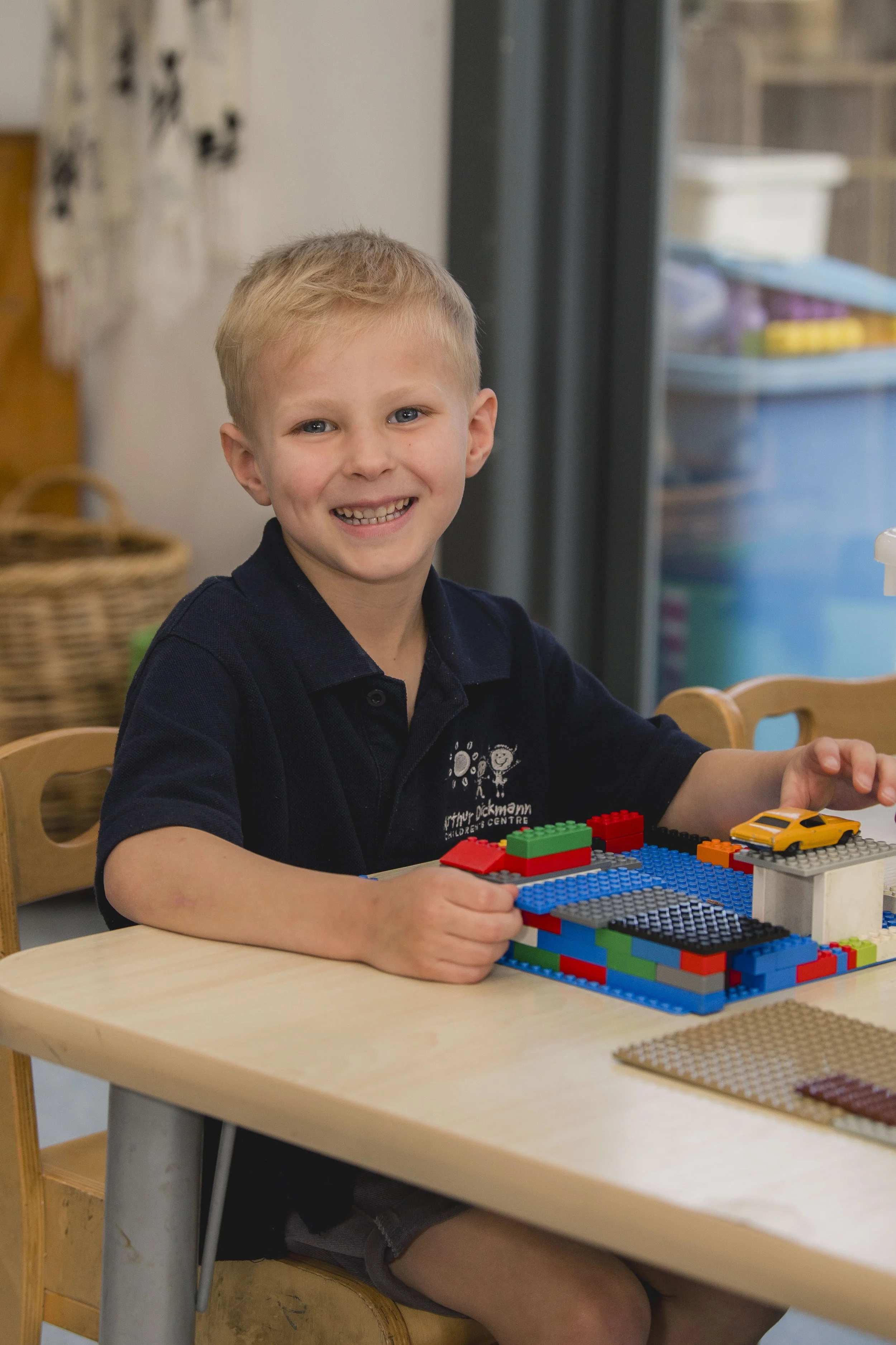 A young boy smiling and sitting at a desk playing with colorful LEGO bricks and a small yellow toy car.