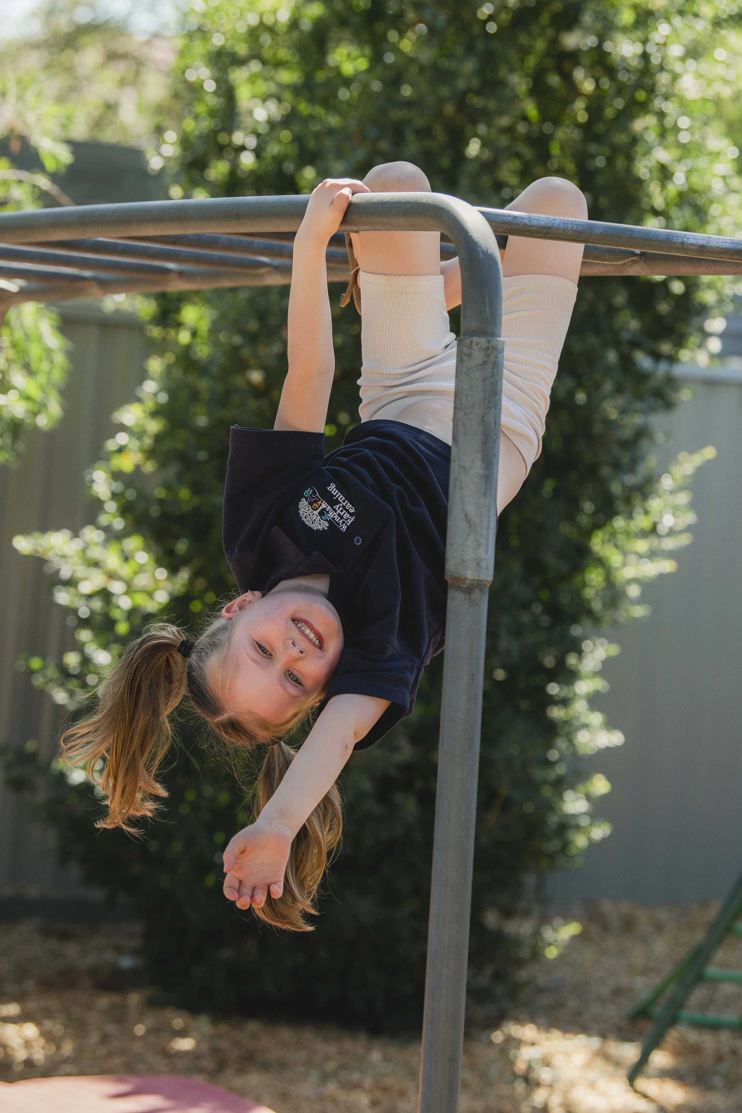 A young girl with long red hair hanging upside down from a jungle gym, smiling at the camera, outdoors with trees and sunlight in the background.