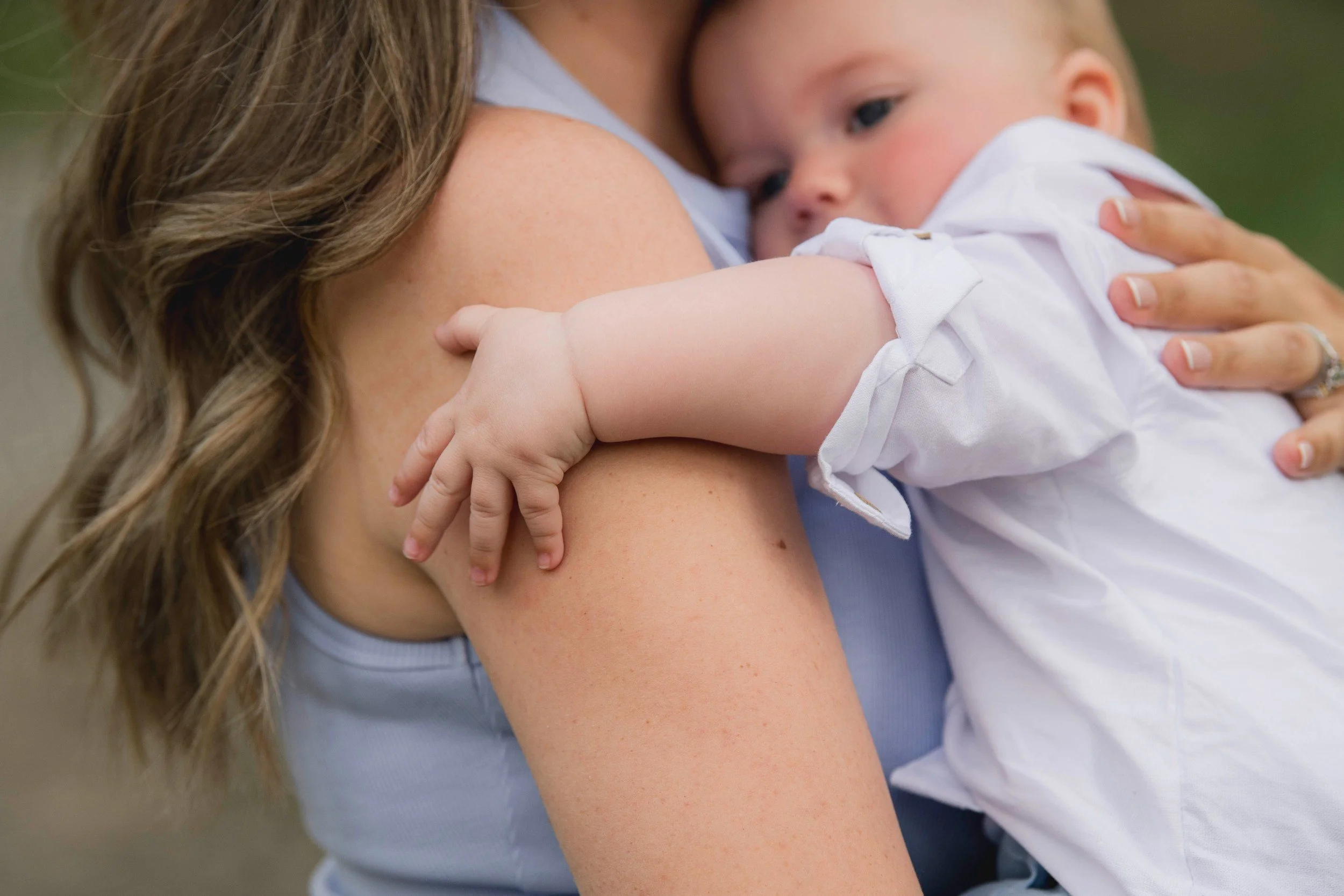 A woman with long wavy hair holding a young boy close, both showing affection. The woman is wearing a light blue shirt and the boy has a short haircut and is wearing a white shirt. The background is blurred with natural outdoor colors.
