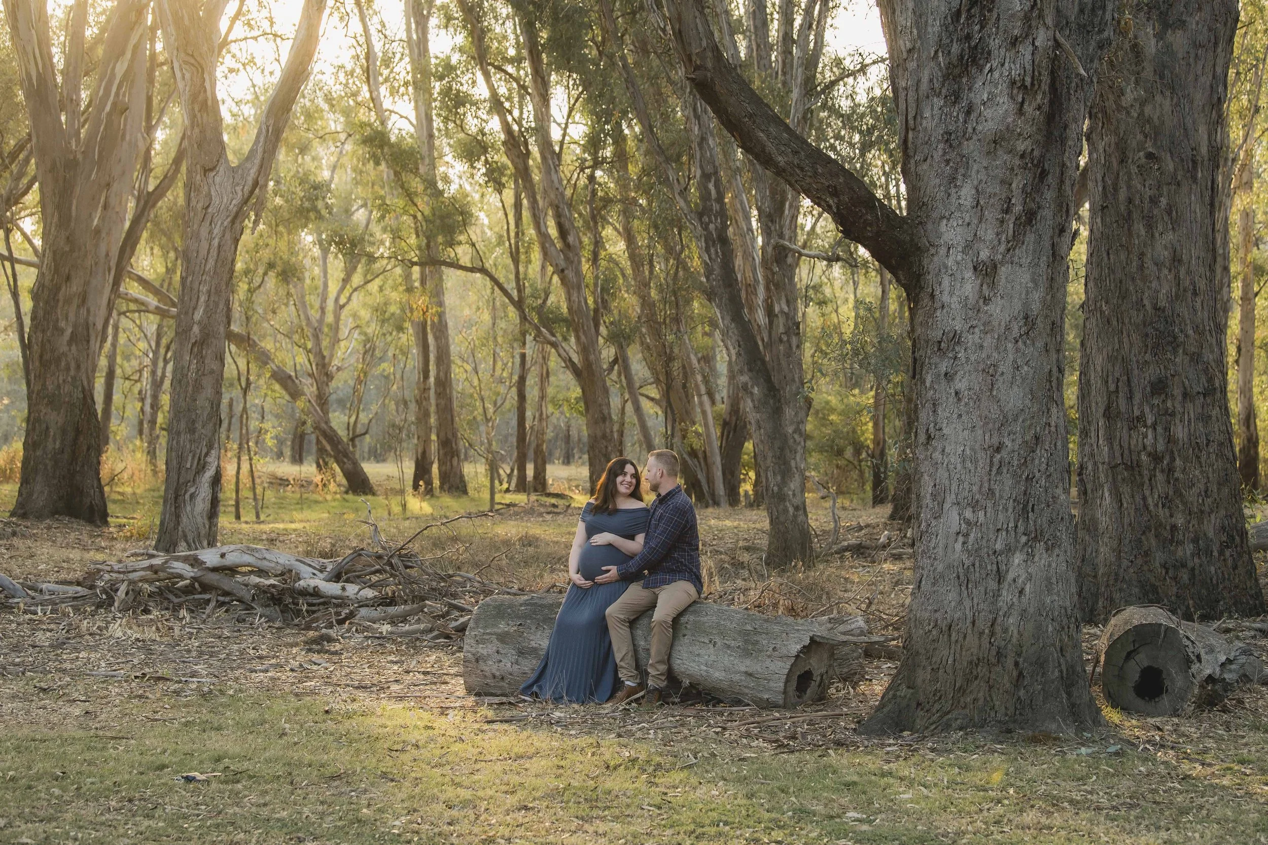 A couple sitting on a large log in a wooded park, with the woman visibly pregnant, sharing a joyful moment during sunset.