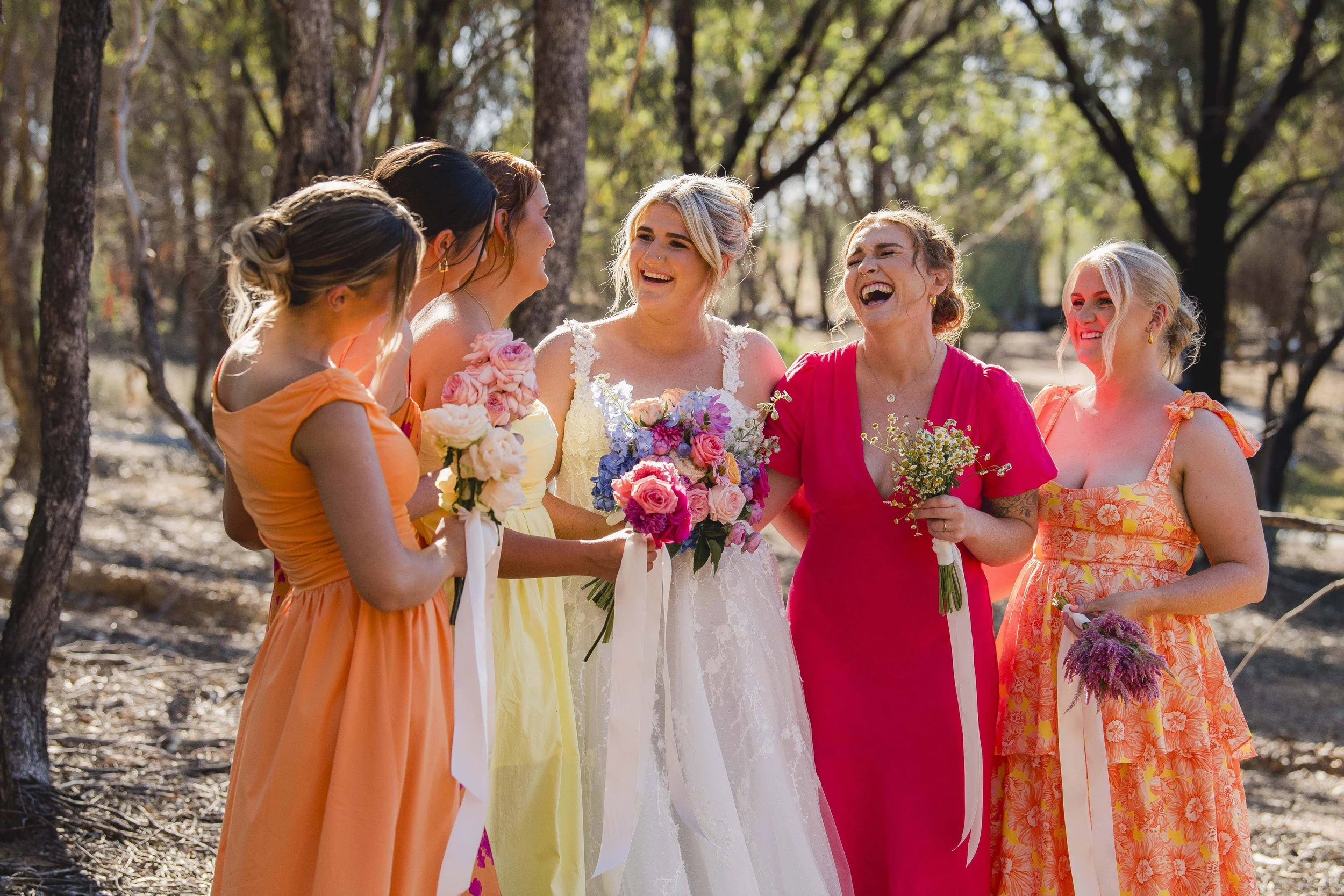 A group of six women dressed in colorful dresses, holding flowers, and laughing together outdoors in a wooded area, celebrating a special occasion.