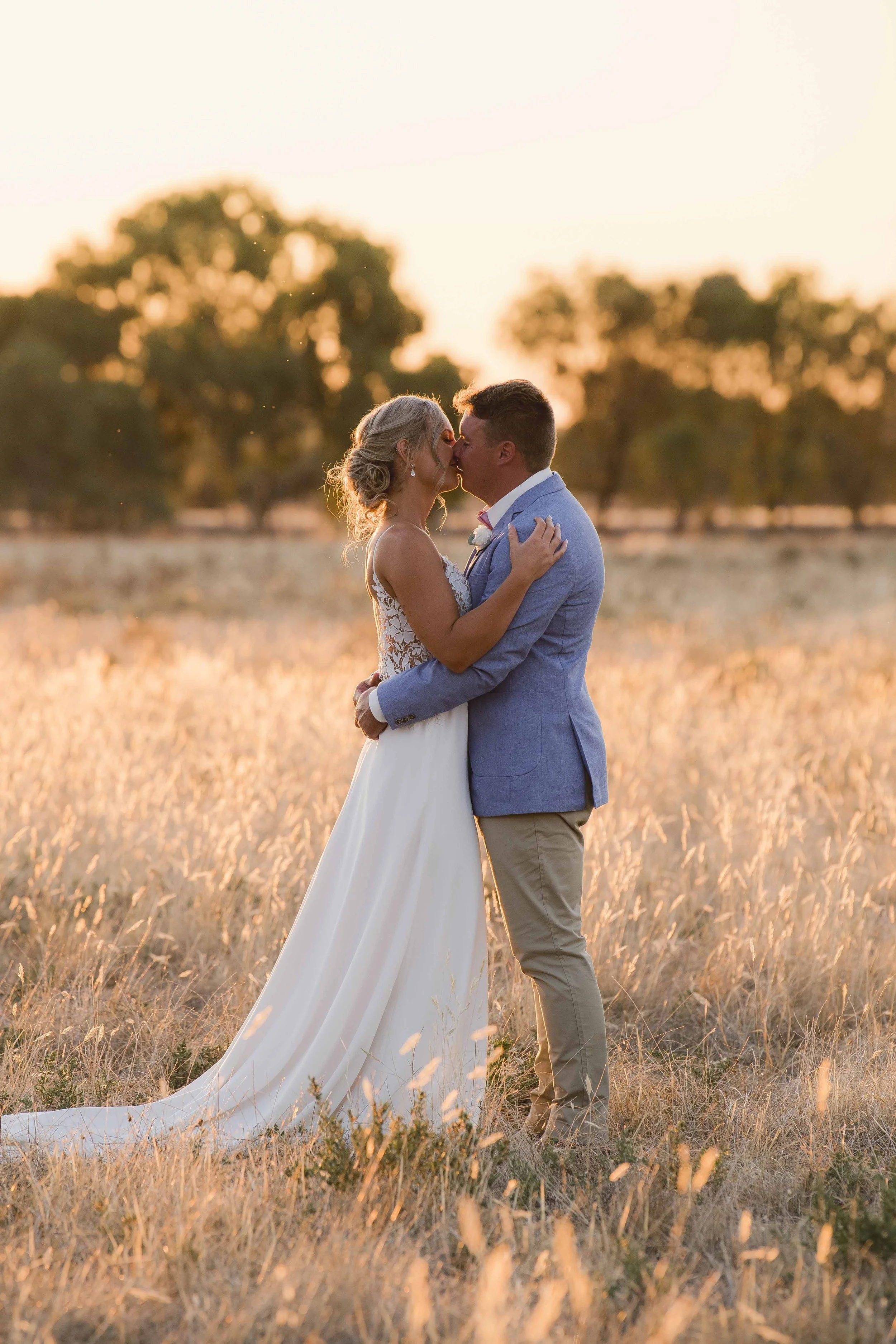 A bride and groom share a romantic kiss in a field at sunset, with trees blurred in the background.