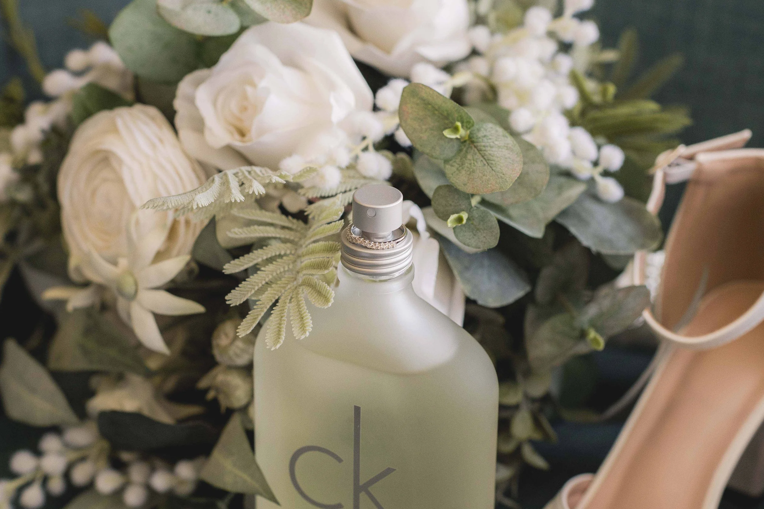 A floral arrangement with white roses, eucalyptus leaves, and other greenery, accompanied by a white bottle with a silver cap, possibly a spray or lotion, and a partial view of a pink cup in the background.