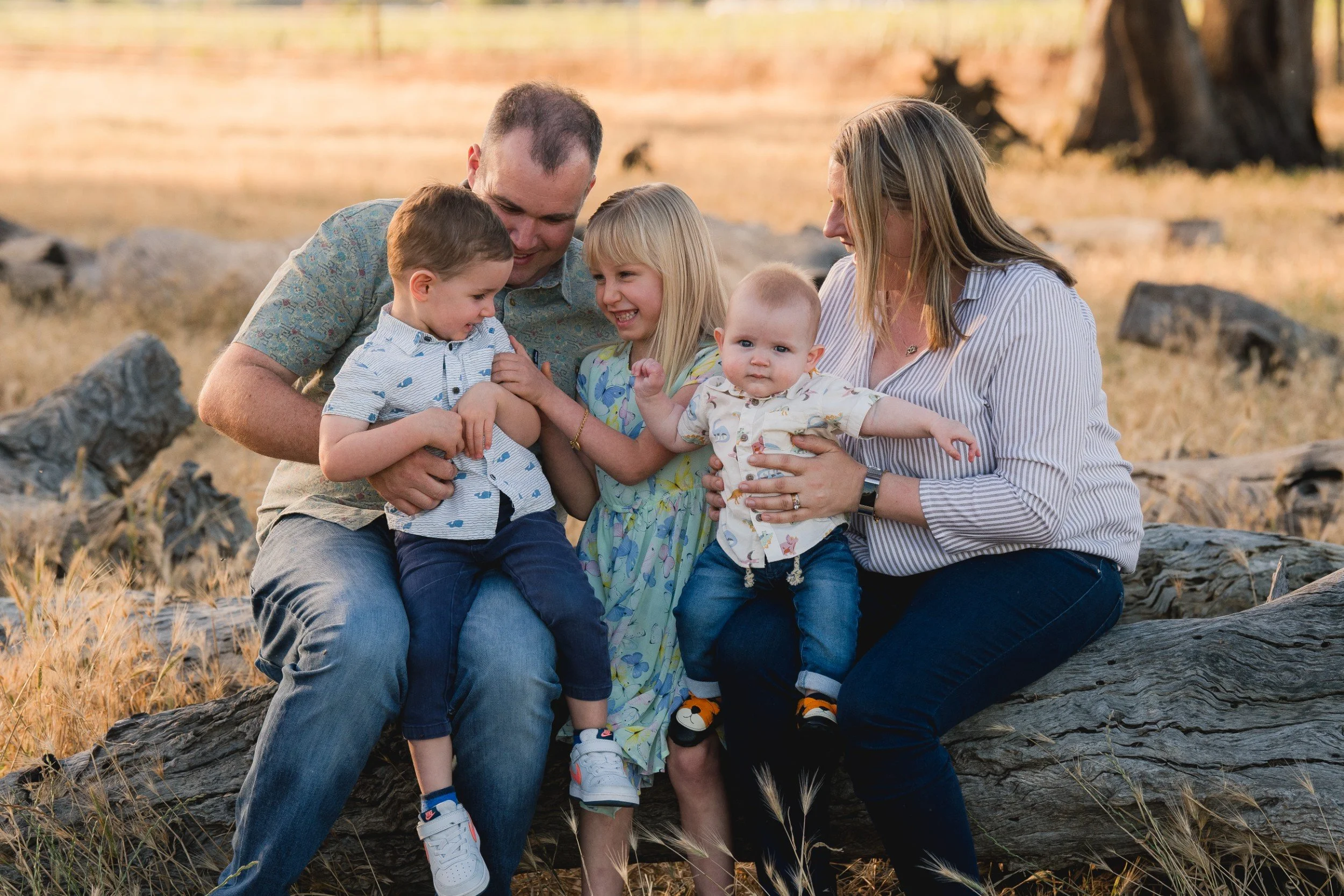 A family of six sitting on a large log outdoors. The father and mother are holding their children, smiling and enjoying a moment together. The children appear to be happy and engaged, with one child reaching out and showing a curious expression. The background suggests they are in a natural setting with trees and dry grass, possibly during sunset.