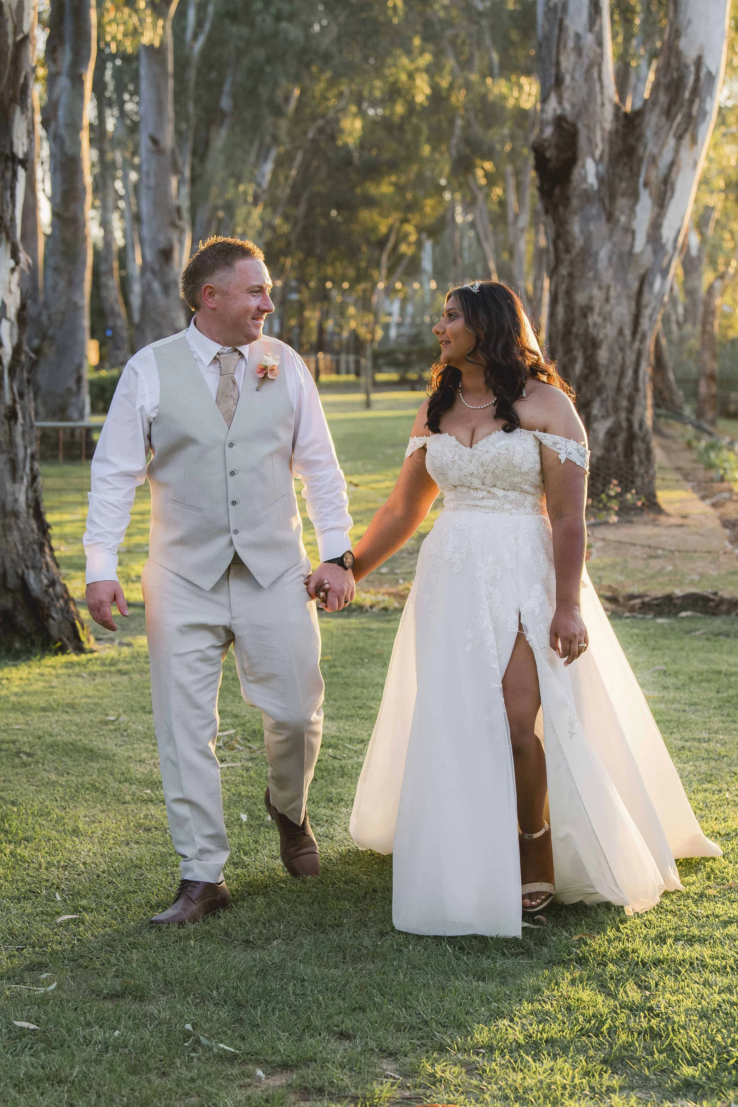 A couple dressed in wedding attire holding hands and walking outdoors in a park-like setting surrounded by tall trees during sunset.