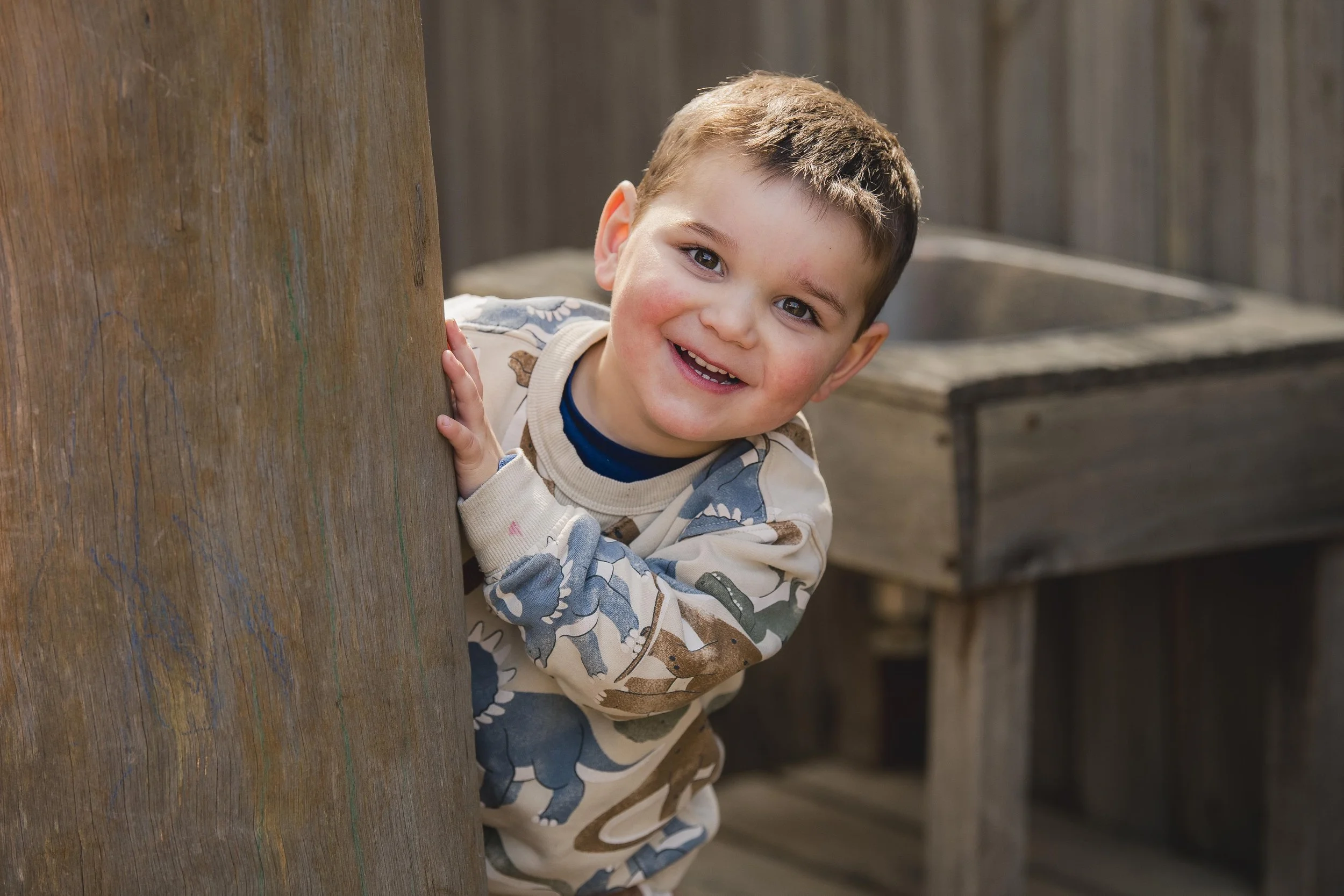 A young boy with short brown hair and gray eyes peeking out from behind a wooden post, smiling, with a wooden fence and a gray container in the background.
