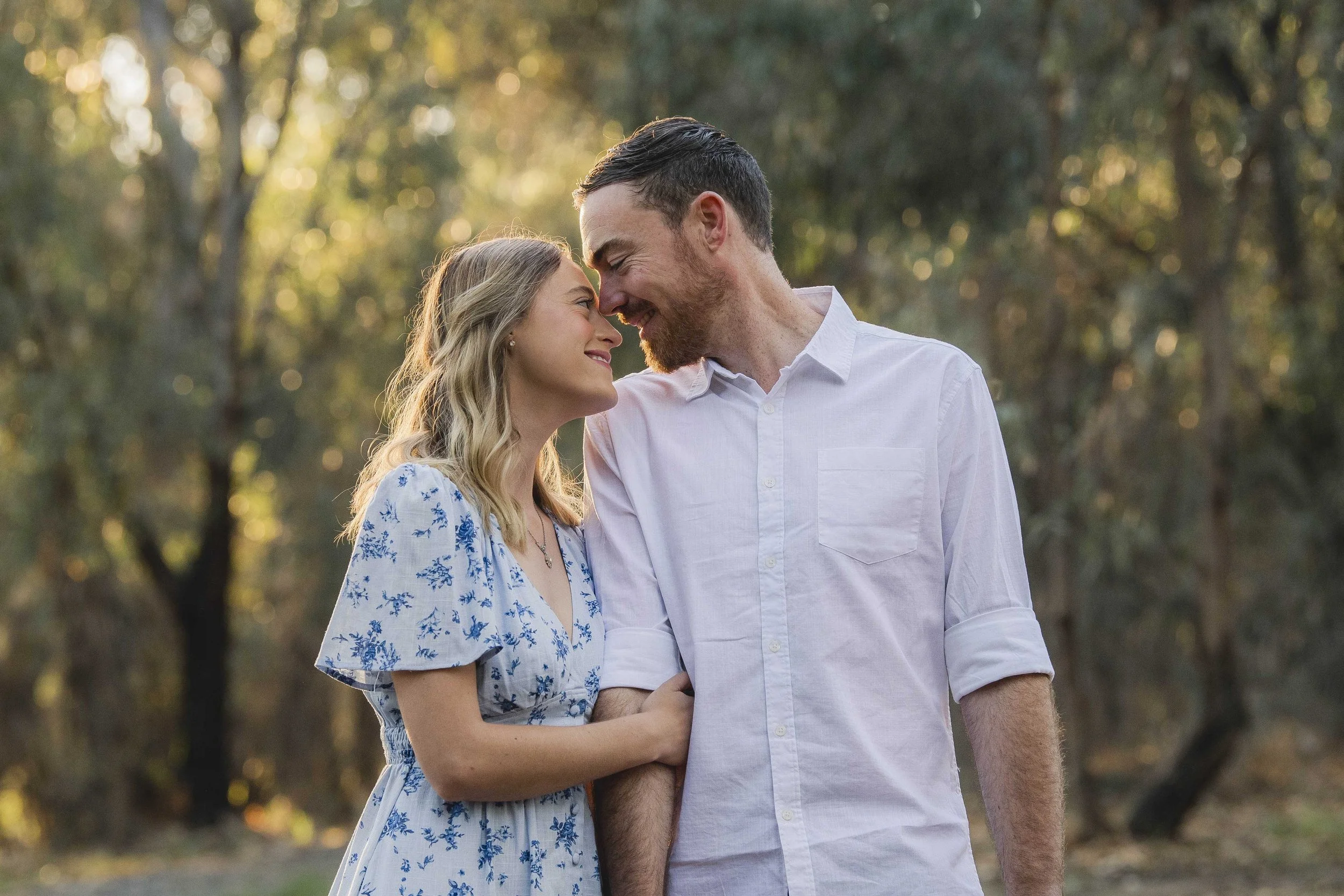 A man and woman are standing close together outdoors, touching foreheads and smiling at each other during sunset in a park or forest with blurred trees in the background.