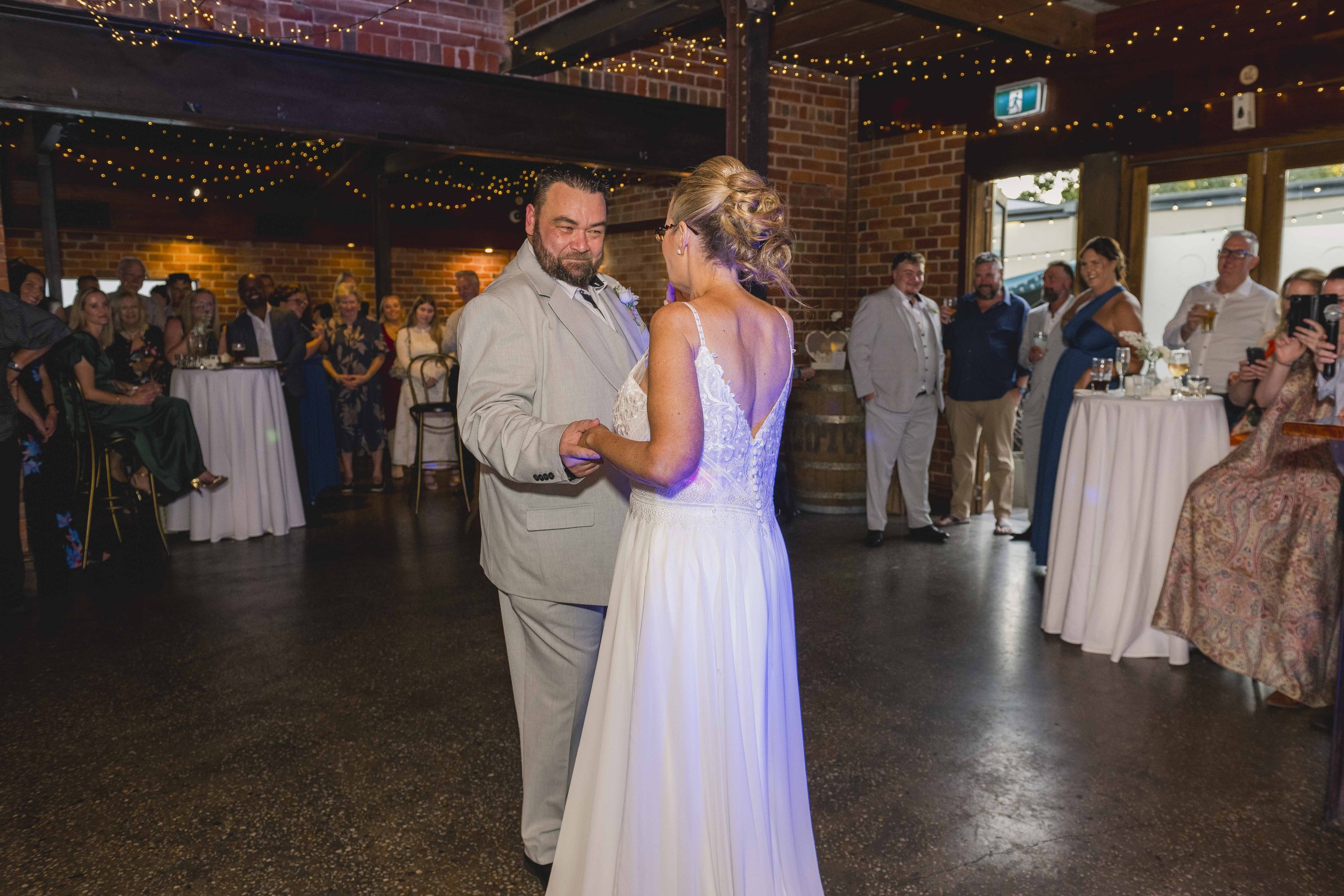 A bride and groom are dancing at their wedding reception, surrounded by guests in a decorated indoor venue with string lights and brick walls.