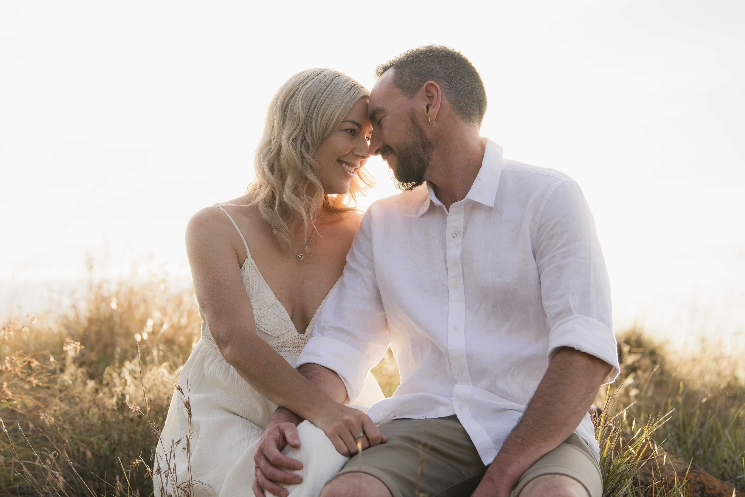 A couple sitting outdoors in a field at sunset, touching foreheads, smiling, and holding hands.