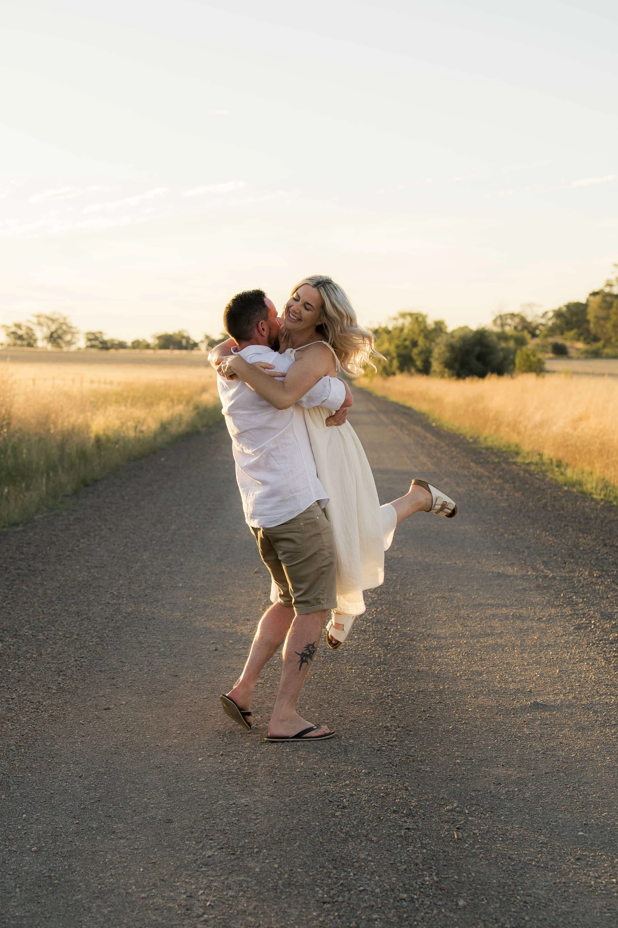 A man is lifting a woman in a white dress on an empty rural road during sunset, both smiling and embracing each other.