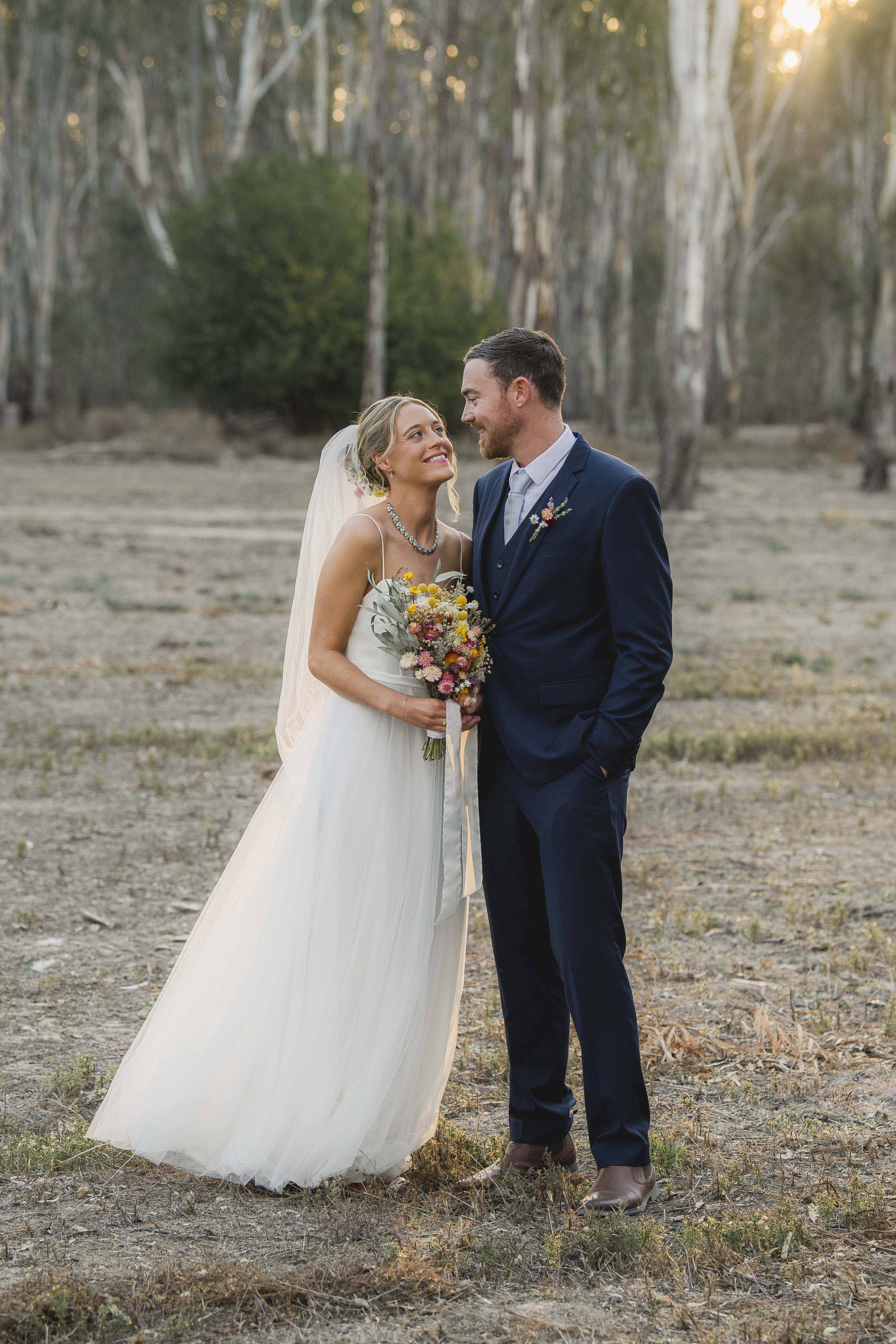 A bride and groom standing in a dirt clearing among trees, smiling at each other during sunset, with the bride holding a colorful bouquet.
