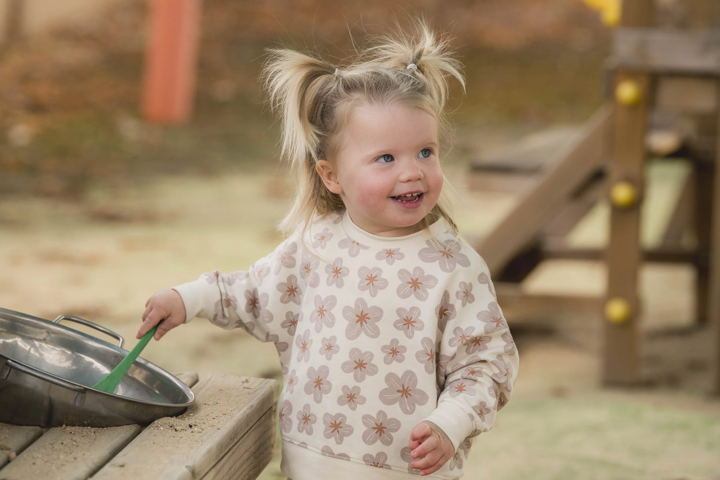 A young girl with blonde hair in pigtails, wearing a long-sleeve shirt with a flower pattern, is outdoors near a sandbox.