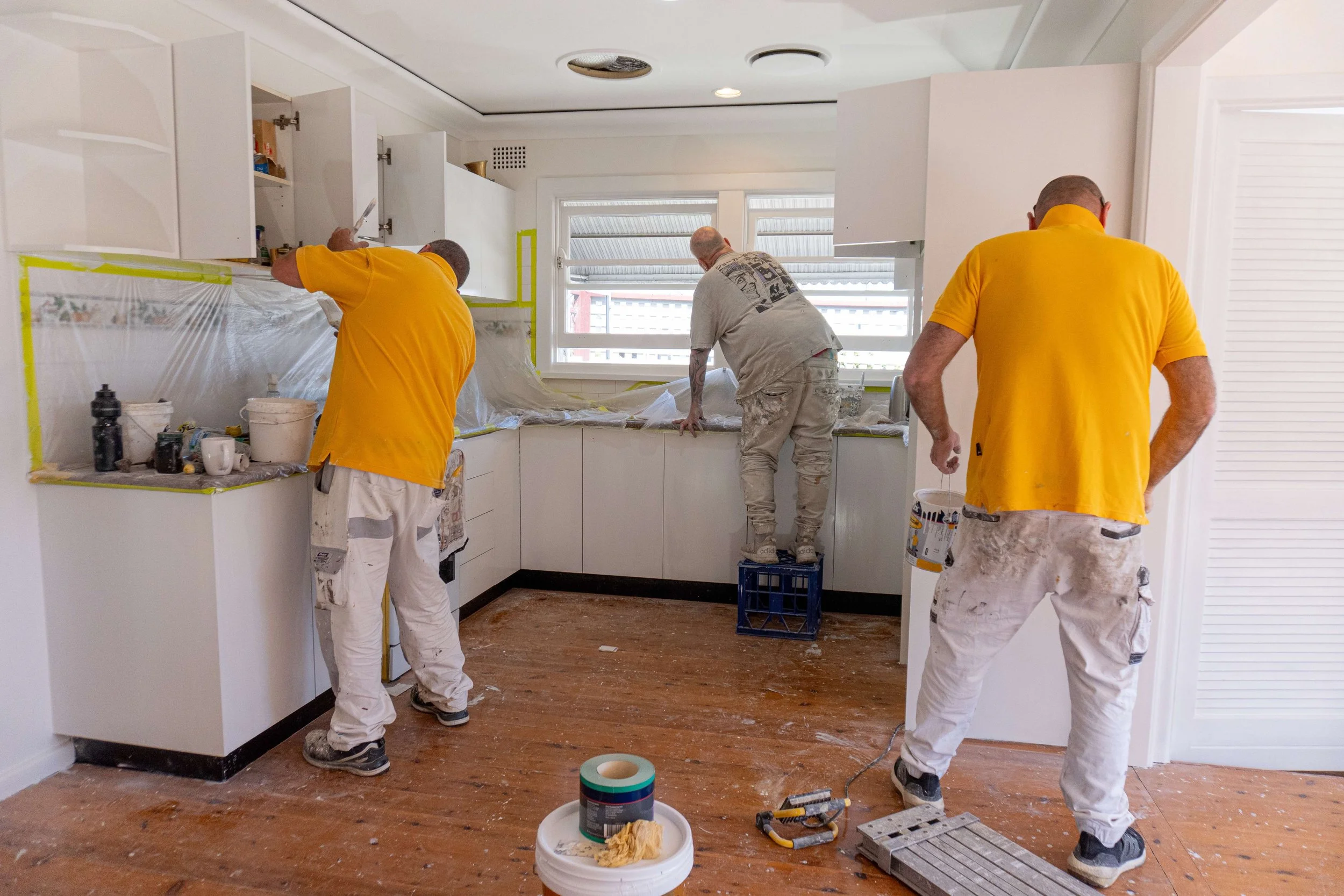 Three workers painting a kitchen, wearing yellow shirts and white overalls, with masking tape around cabinets and plastic covering surfaces.