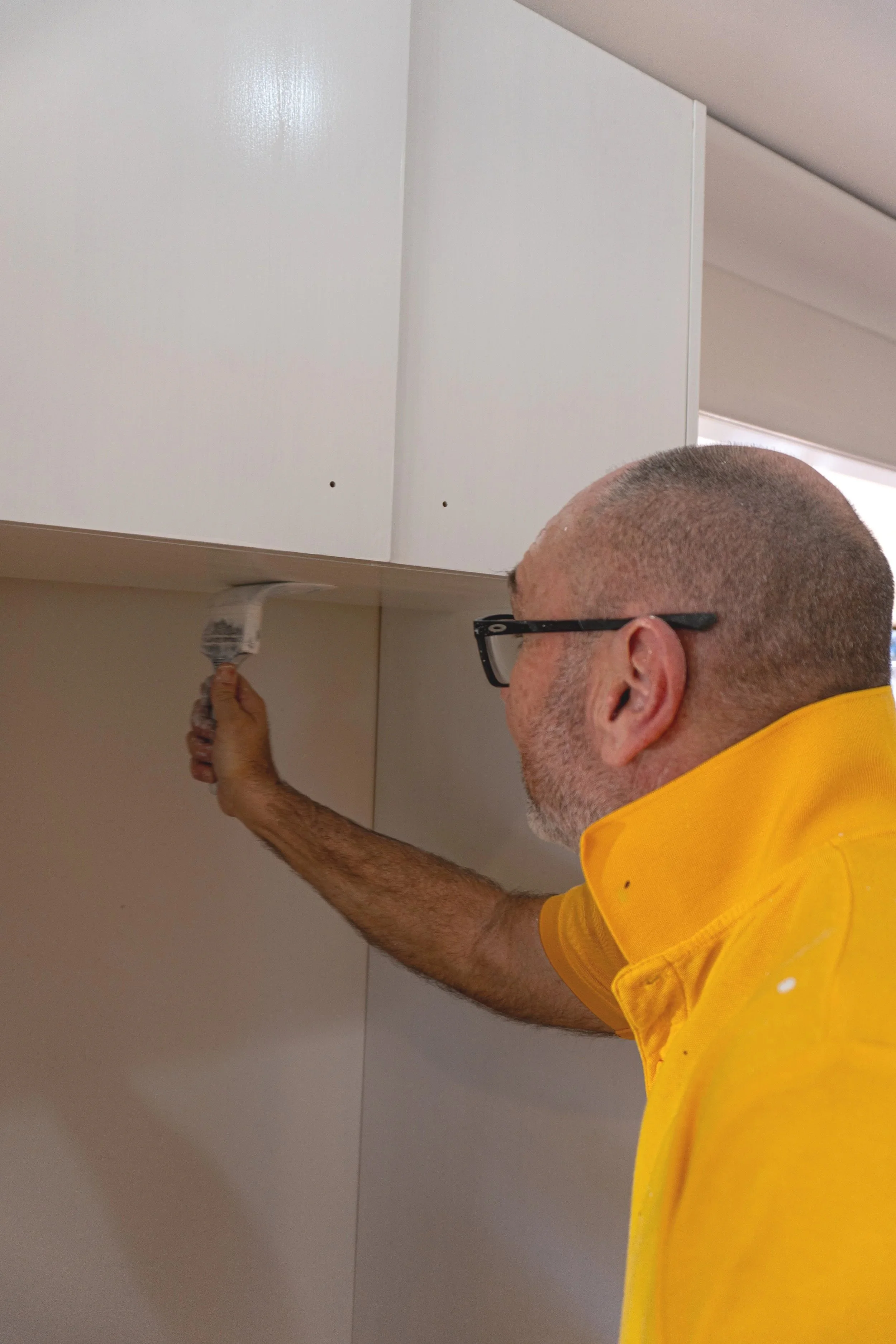 A man with glasses and a beard painting the underside of a white kitchen cabinet with a paintbrush.