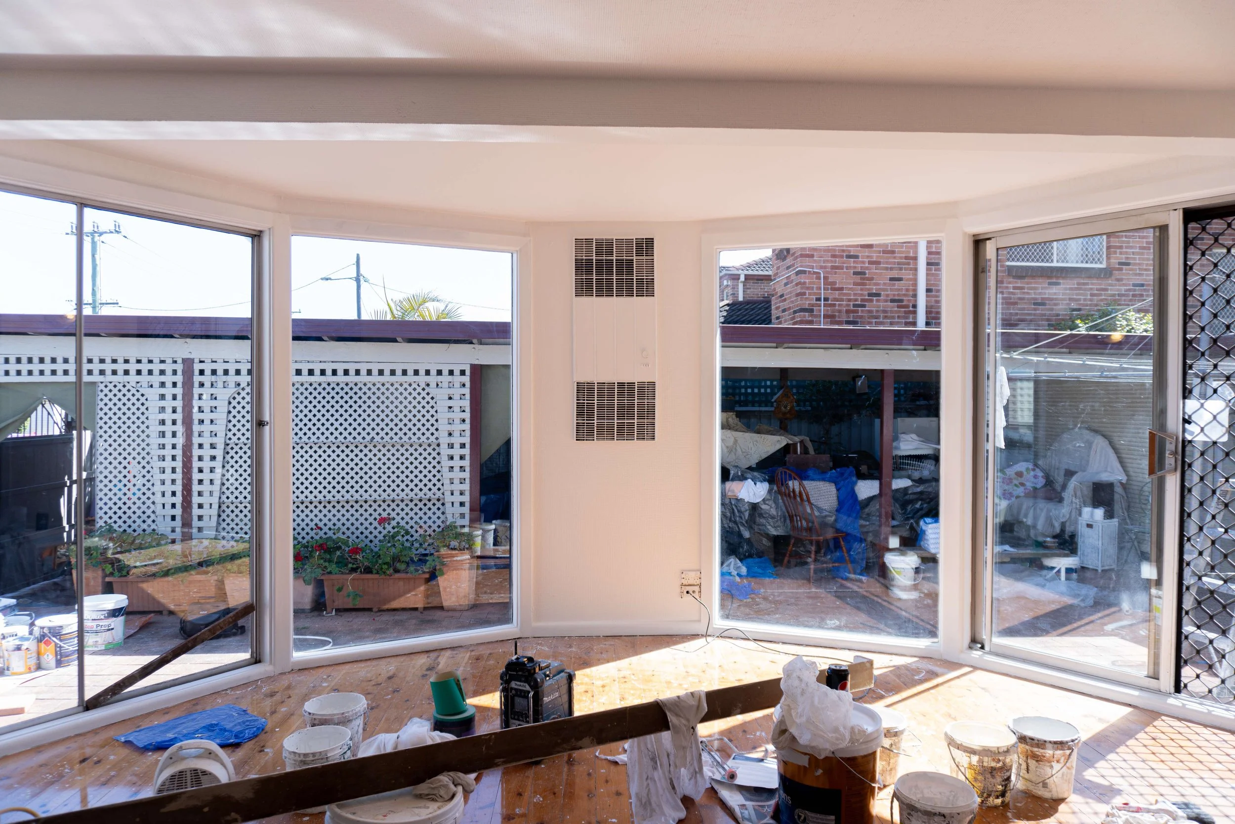 Interior of a house with large sliding glass doors leading to an outdoor patio, which has garden planters, construction tools, and painting supplies scattered inside and outside.