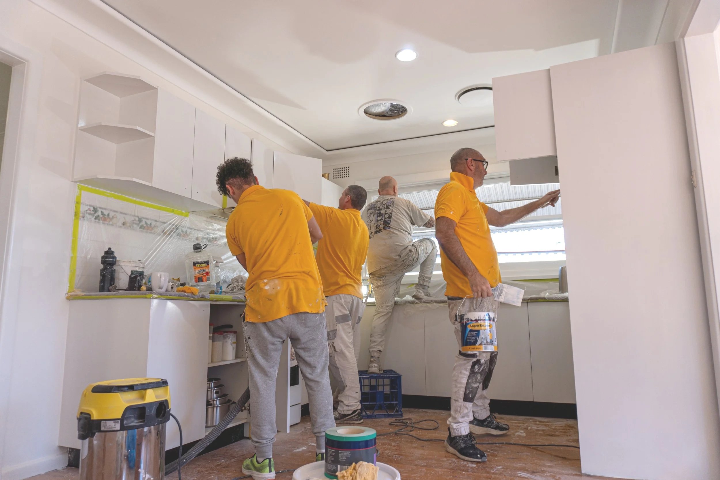 Five men painting and working on kitchen cabinets in a bright kitchen, with painting supplies and tools on the counter and floor.