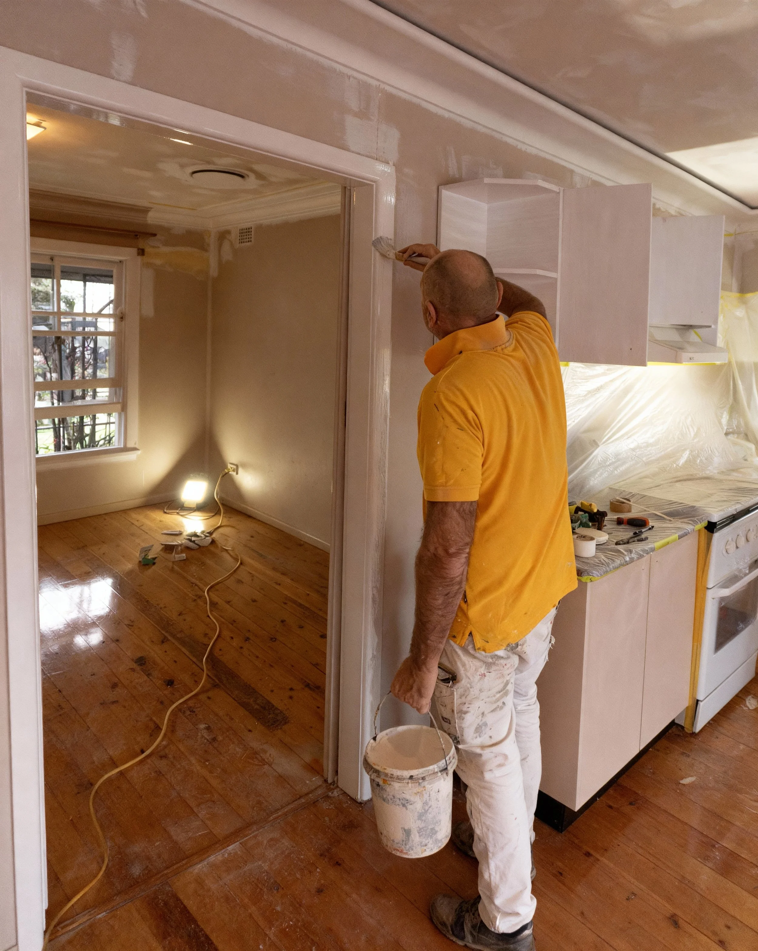 A man wearing a yellow shirt and white pants painting the trim of an interior wall in a home renovation project.