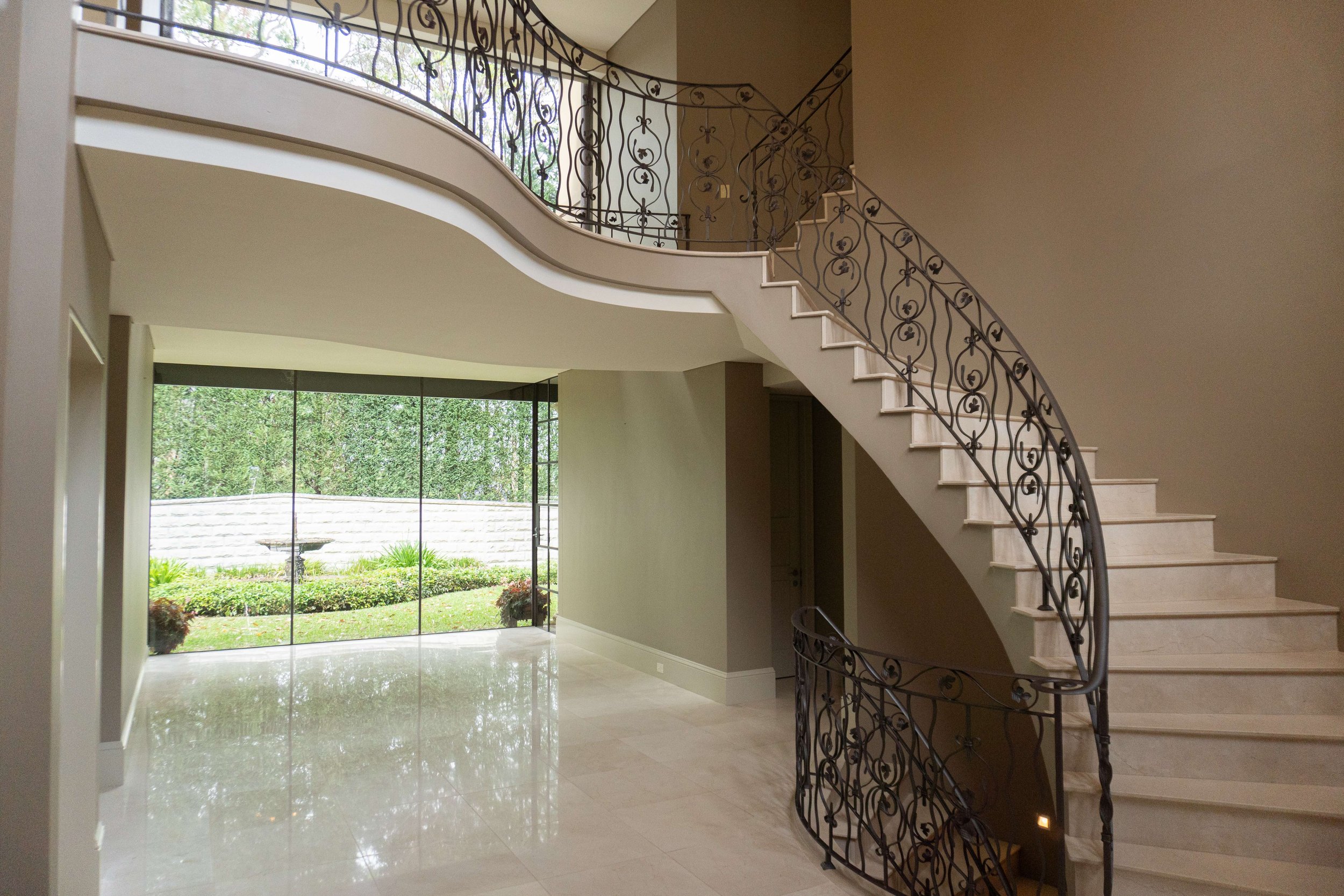 Interior view of a spacious foyer with a curved staircase featuring decorative black wrought iron railing, large floor-to-ceiling window showing garden outside, and polished light-colored tile flooring.