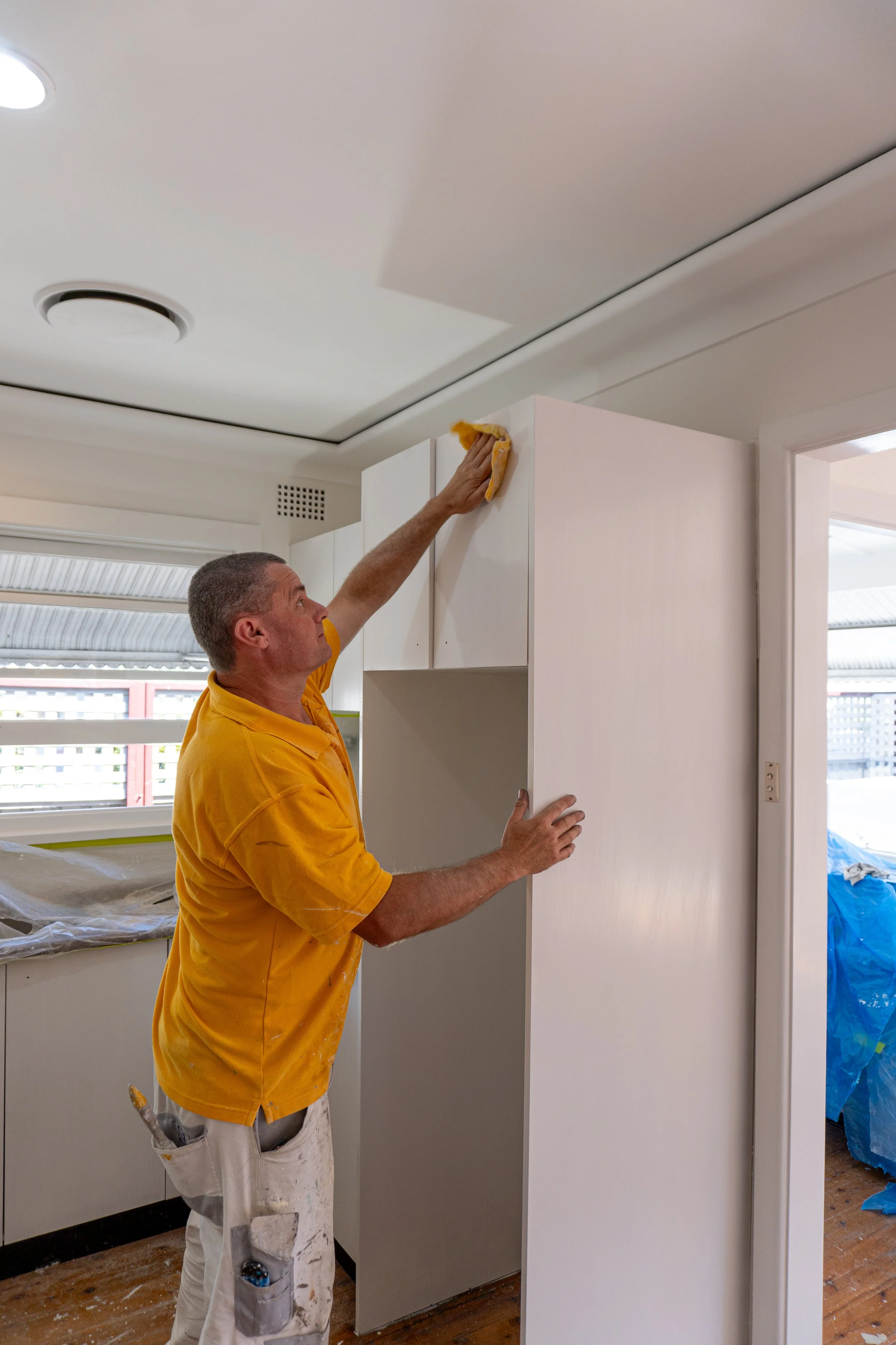 A man in a yellow shirt is cleaning a white cabinet with a cloth in a room that appears to be under renovation.