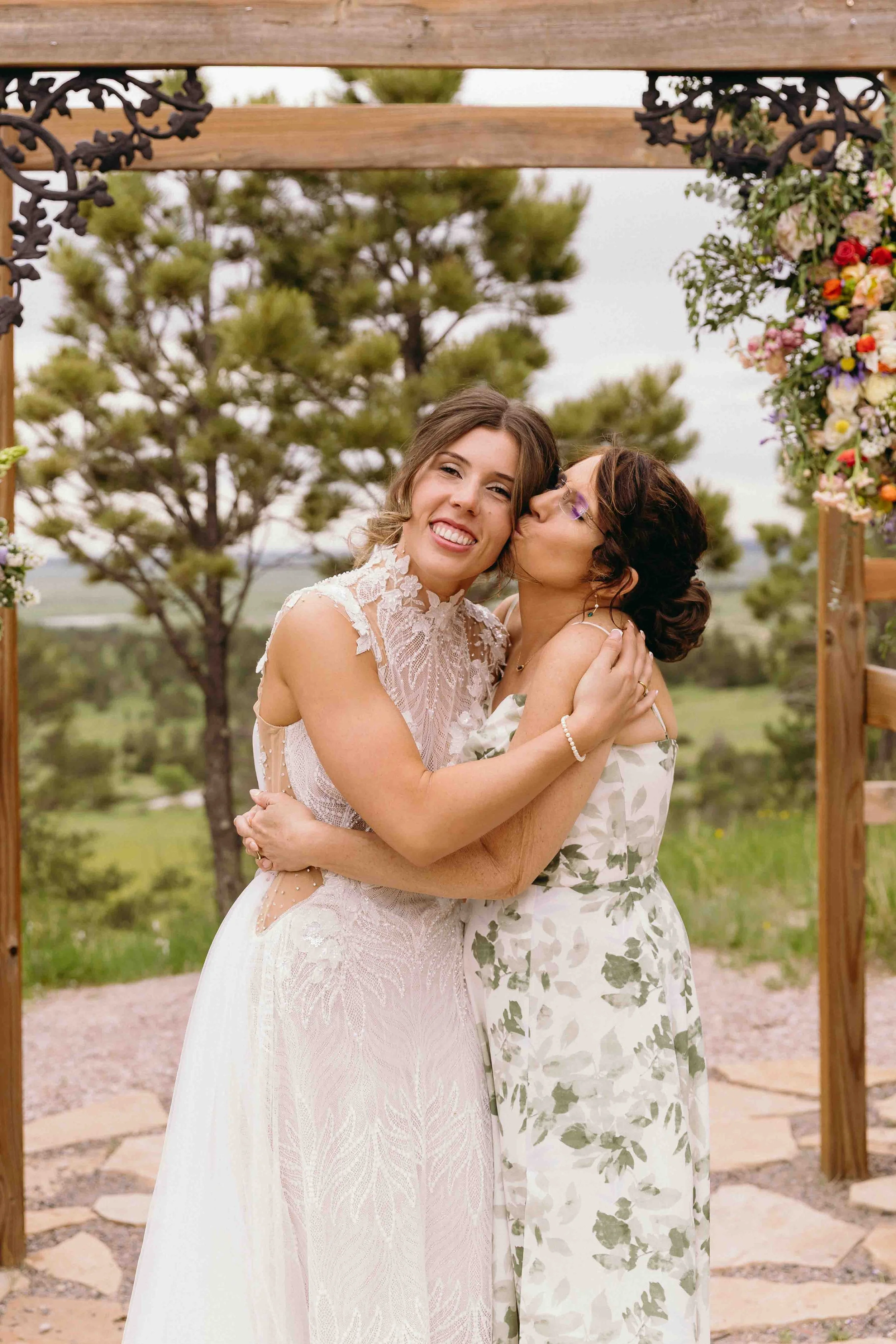 Two women hugging and sharing a kiss at an outdoor wedding ceremony with trees and flowers in the background.