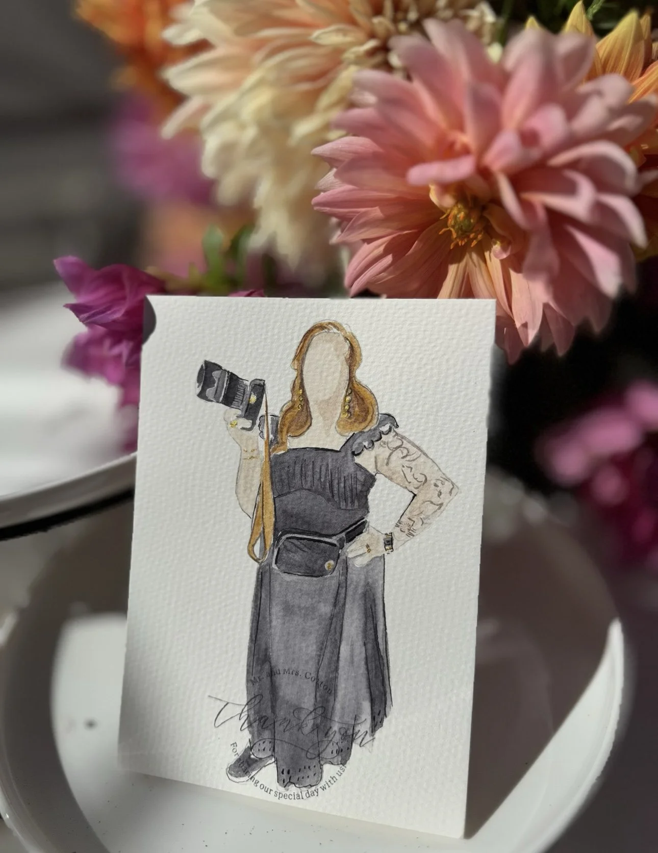 A watercolor illustration of a woman with long red hair, tattoos on her arms, wearing a black dress with puffy sleeves, holding a camera in one hand, and standing in front of pink and cream flowers.