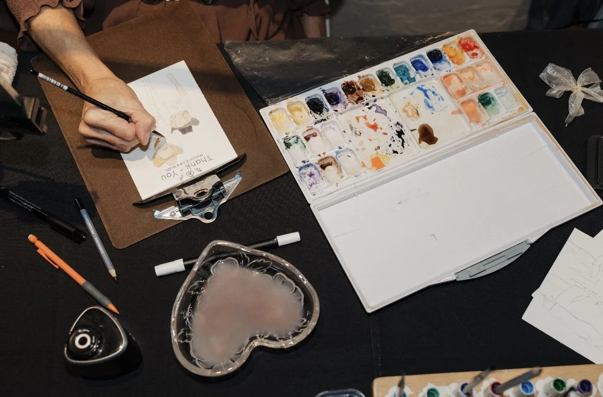 Person painting a picture of a woman with watercolor paints on a table with art supplies, including a palette, brushes, pens, and paper.