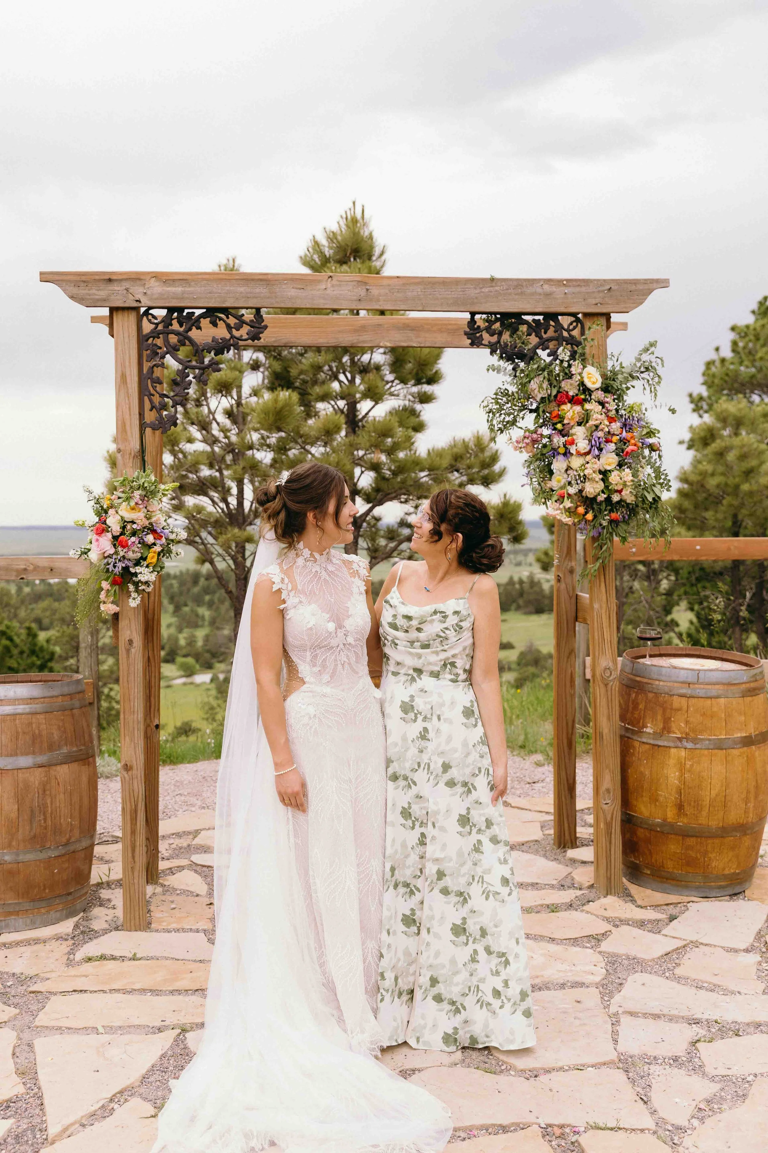 Two women in wedding and formal dress standing under a decorated wooden arch outdoors, holding hands and smiling at each other, with trees and a cloudy sky in the background.