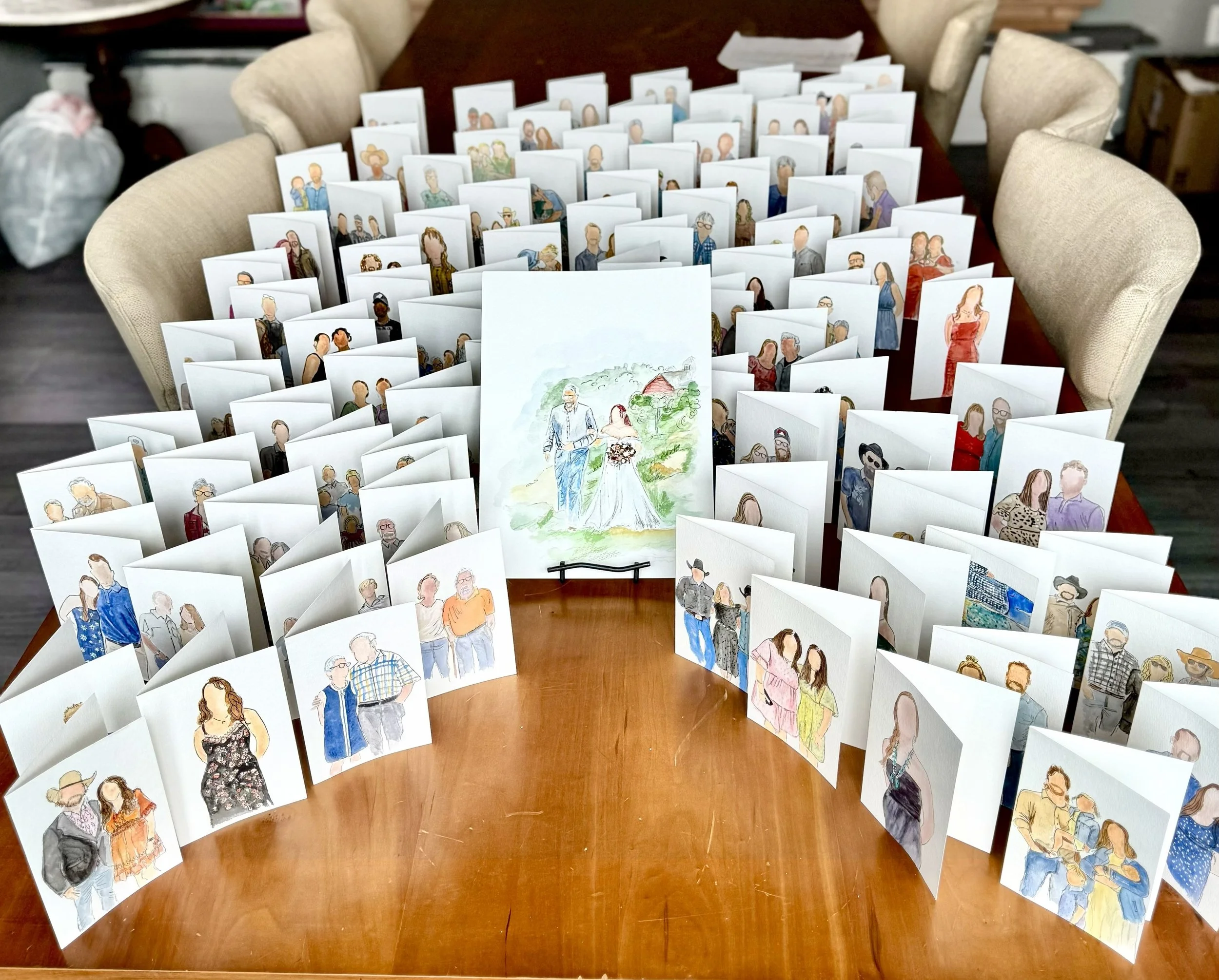 Numerous handmade wedding cards with watercolor illustrations of couples, arranged on a large wooden table, with a centerpiece illustration of a bride and groom walking in a countryside setting.