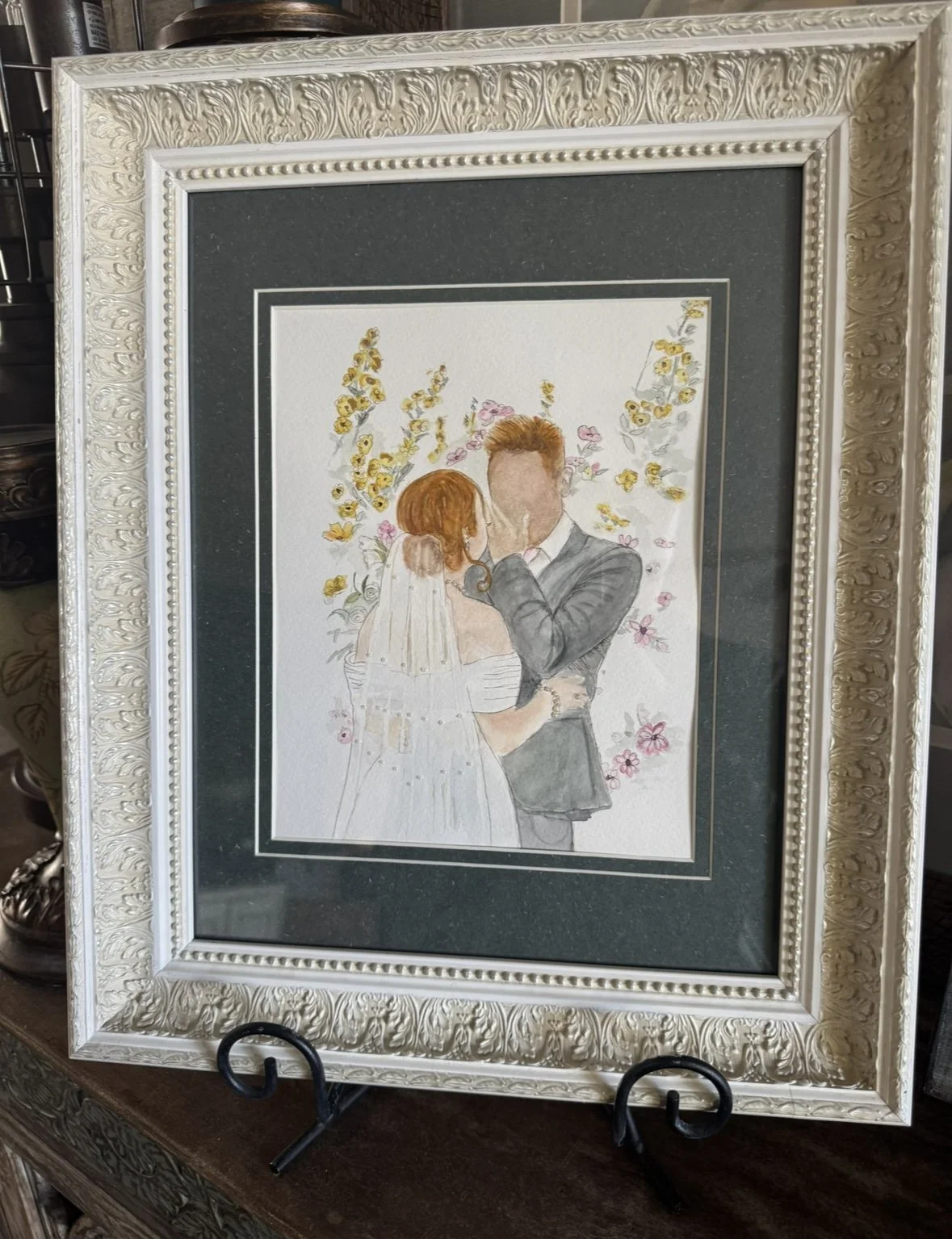 A framed watercolor painting of a bride and groom sharing a kiss, surrounded by colorful flowers, with an ornate silver-colored frame and a dark gray mat.