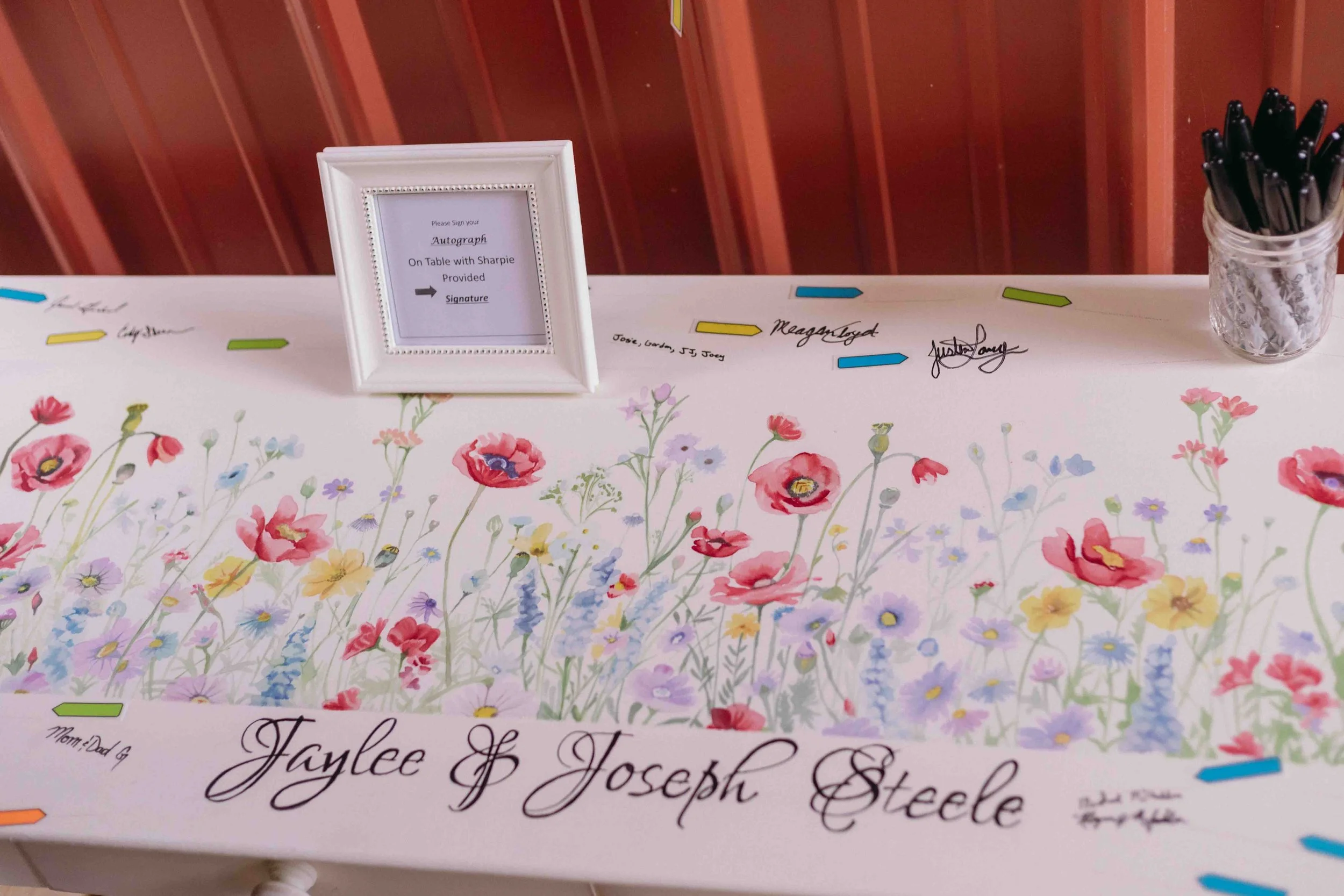 A decorated guestbook table with a floral cover, featuring signatures and messages, a framed sign instructing guests to sign with a Sharpie, and a container filled with black markers. The guestbook is personalized with the names Jaylee and Joseph Steele.