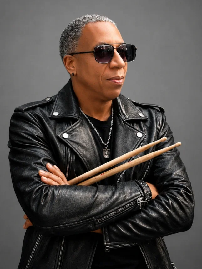 A man with short gray curly hair wearing sunglasses, a black leather jacket, and a chain necklace, crossing his arms and holding drumsticks, against a gray background.