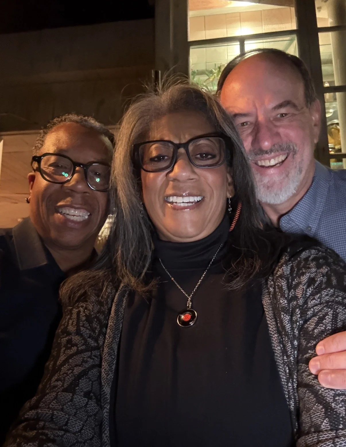 Tim Hutson on the left, Anita Guris in center and Paul Guris on right.  Smmiling and taking a selfie together indoors, near a window with a warm ambient light at SOUTH Jazz Club
