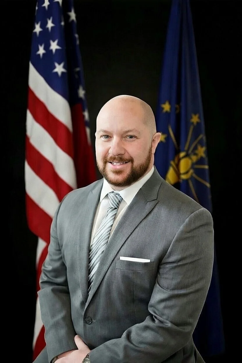 A man in a gray suit, white shirt, and gray striped tie standing in front of American and Indiana state flags.