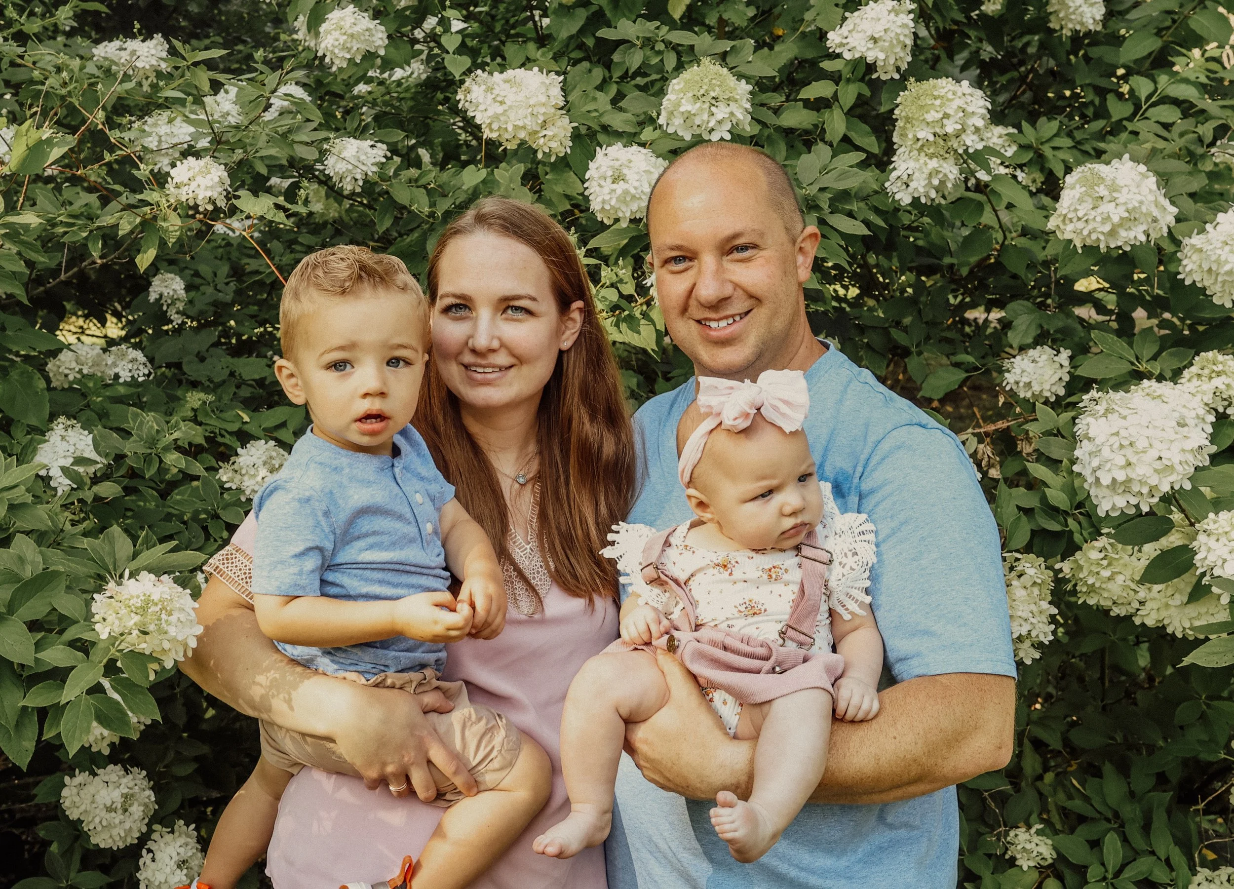 A family of four outdoors beside a leafy bush with white flowers. The mother has long brown hair, a pink top, and holds her young son. The father has a shaved head, a blue shirt, and holds a young girl with a pink bow headband. All are smiling.