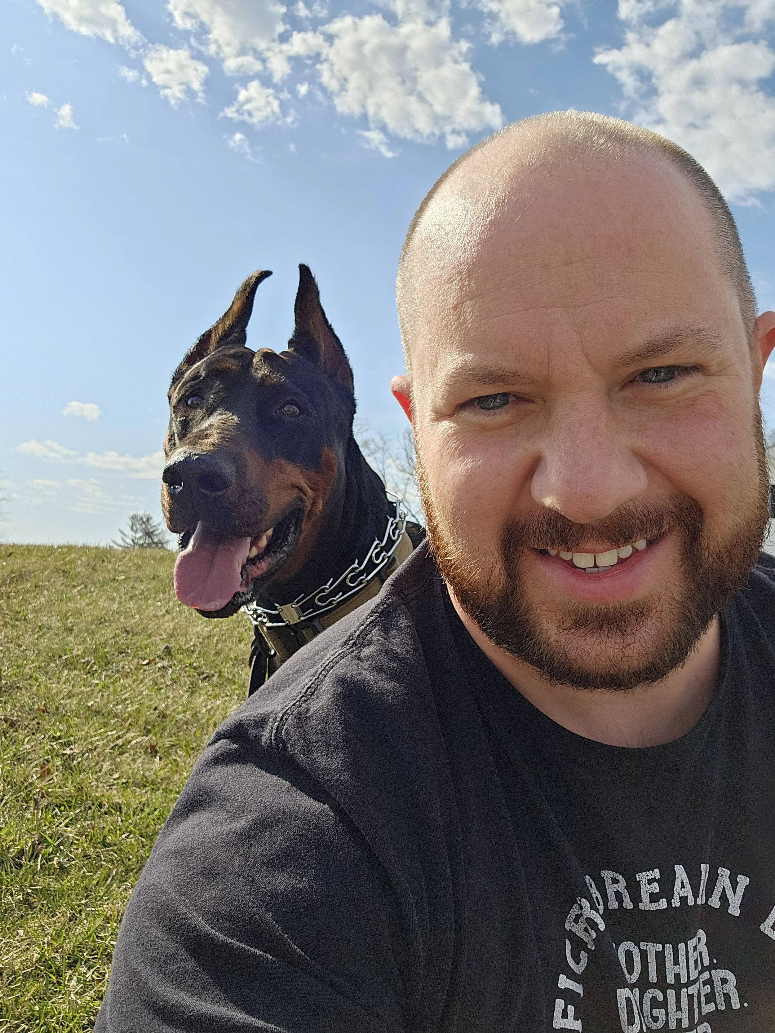 A man with a beard and a black T-shirt taking a selfie outdoors on a sunny day, with a Doberman dog behind him on the grass, clear blue sky with some clouds.